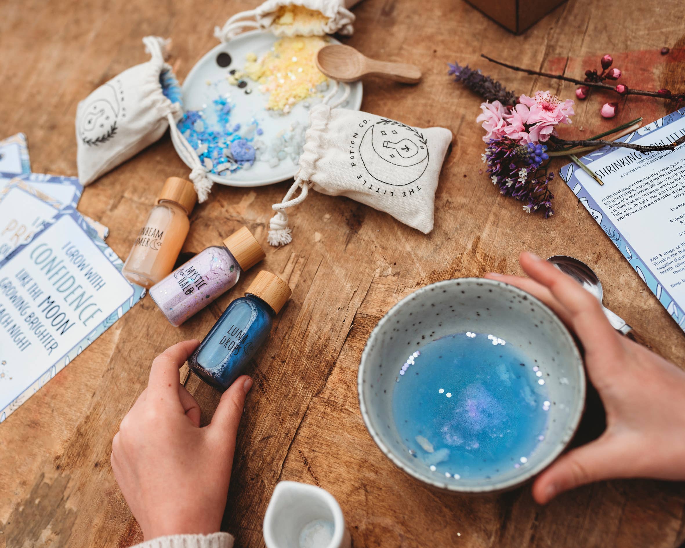 Close-up of a wooden table with a DIY craft setup. A person holds a small bottle of blue dye over a bowl of blue liquid. Various small pouches, dried flowers, and spoons are spread out alongside instruction cards labeled "Confidence Dance" and "Pink Moon," creating an enchanting Moon Magic MINDFUL Magic Potion Kit by THE LITTLE POTION CO, perfect for mindfulness activities.