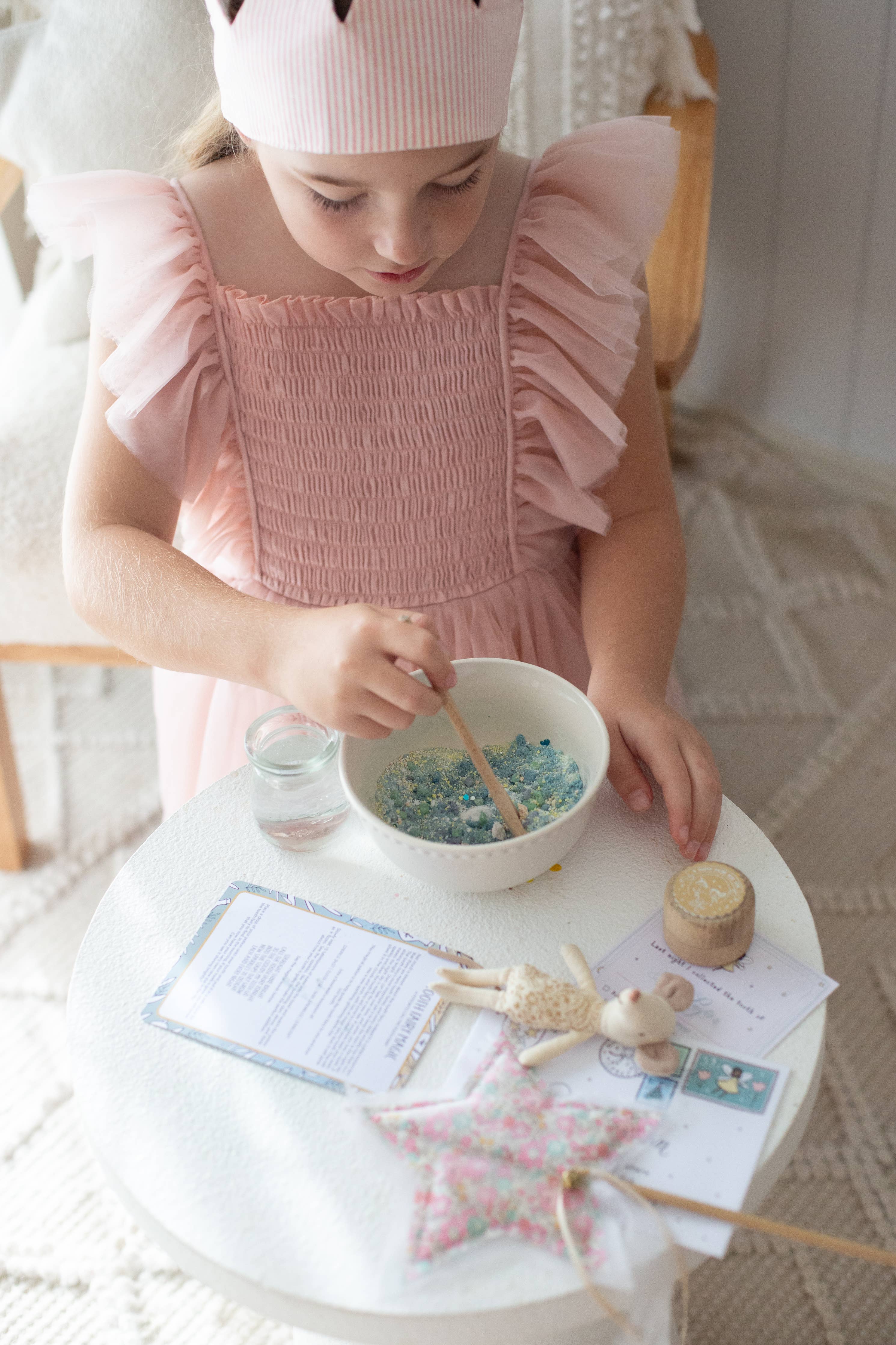 A child wearing a pink dress and hat stirs the magical Tooth Fairy Magic Potion Pouch by THE LITTLE POTION CO in a bowl on a small white table. Nearby, sparkly ingredients, craft supplies, a glass of water, paper, and decorative items add to the enchanting scene in the cozy room with its woven rug.