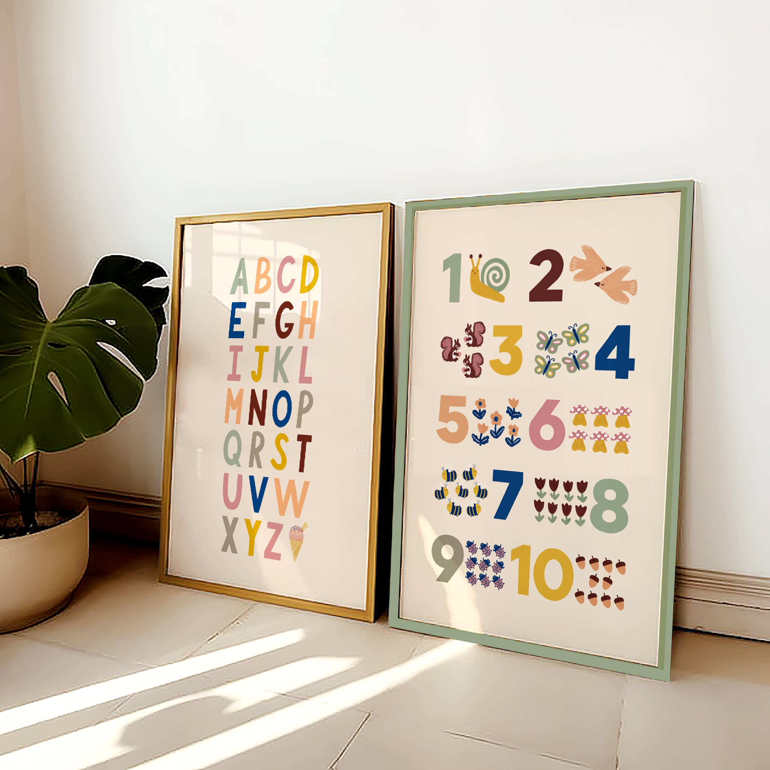 Two framed posters rest against a wall. The left poster, the Bold A-Z Alphabet Wall Art Print by Lauren Sissons Studio, displays an alphabet print in multicolored letters, perfect nursery decor. The right poster features numbers 1 to 10 with corresponding illustrations: snail for 1, doves for 2, butterflies for 3, etc. A large green plant is to the left.