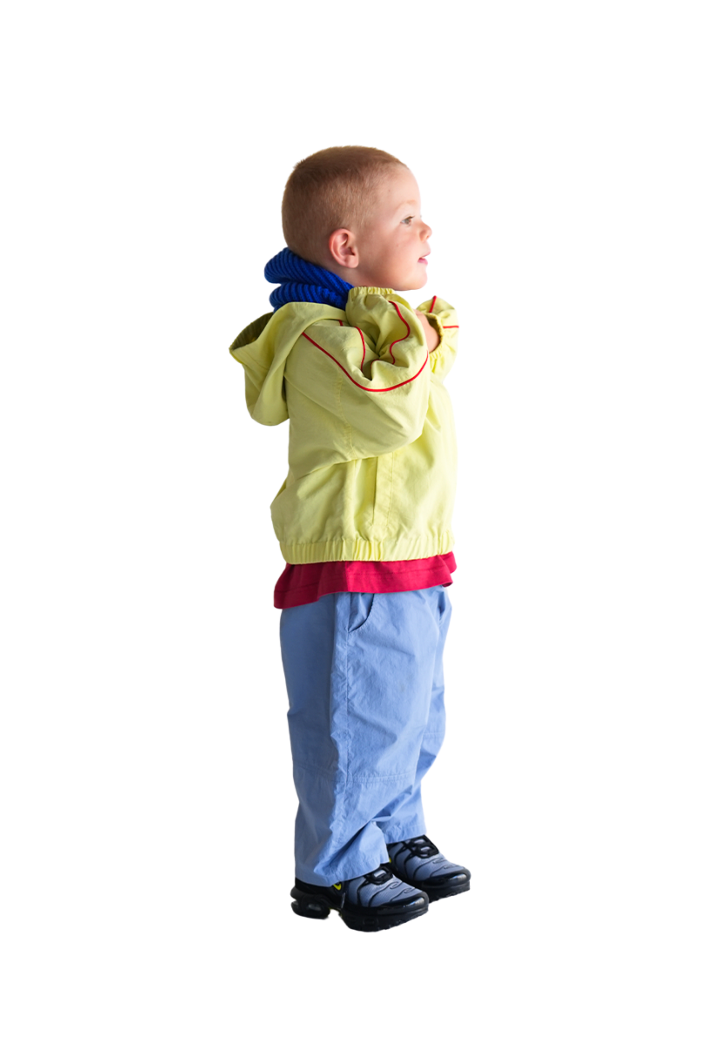 A young child with short hair, wearing the oversized Franco's Dad ~ Tech Spray Jacket Butter by FRANCO'S DAD, a red shirt, blue pants, and black shoes, stands with arms crossed and looks upwards to the left against a white background.