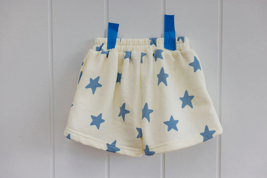 A pair of SUNDAY SIBLINGS ~ Kiddo Shorts Blue, crafted from soft French terry cotton with a playful blue star pattern, are displayed on a white wall using two pieces of blue tape.