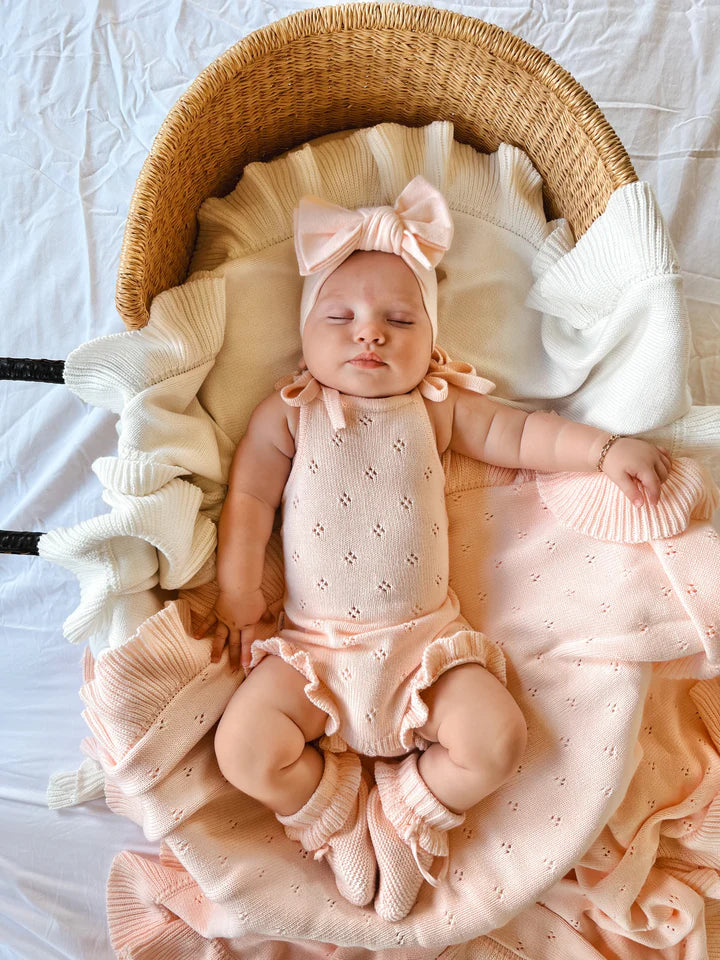 A sleeping baby is lying in a woven basket, lined with white fabric and a ruffled pink blanket. The baby is dressed in ZIGGY LOU's Frill Bodysuit Primrose, a sleeveless, 100% cotton pink outfit from the Primrose Signature Collection, complete with matching leg warmers and a headband with a bow. The background is a white, textured cloth.