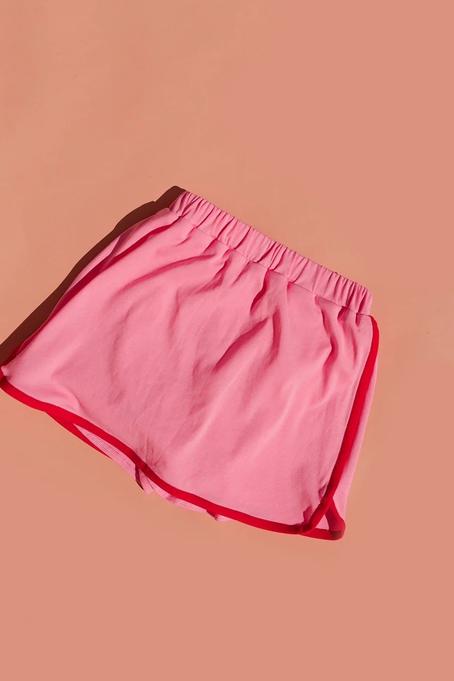 The SMALL SWIM CLUB 70's Skort in pink/red, available in size 7-8, is showcased on a peach background. This retro-designed skort features red trim, an elastic waistband, and a slightly overlapping dolphin hem. Made from 100% cotton for comfort.