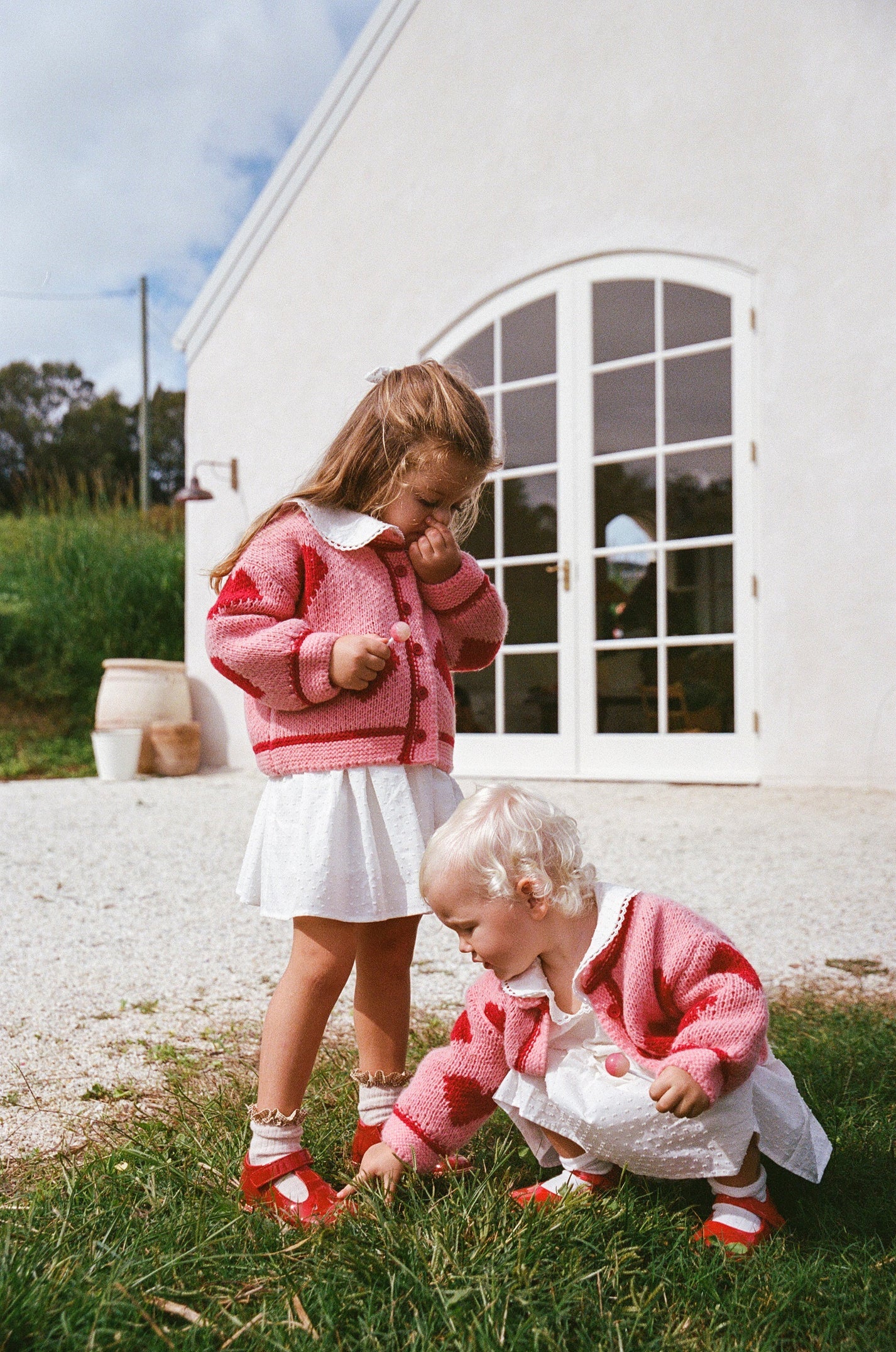 Two young children wear matching The Wholesome Store - Lover Knit Cardigan Mini by THE WHOLESOME STORE over white outfits outside a white building. One stands while the blond-haired child crouches to touch the grass.