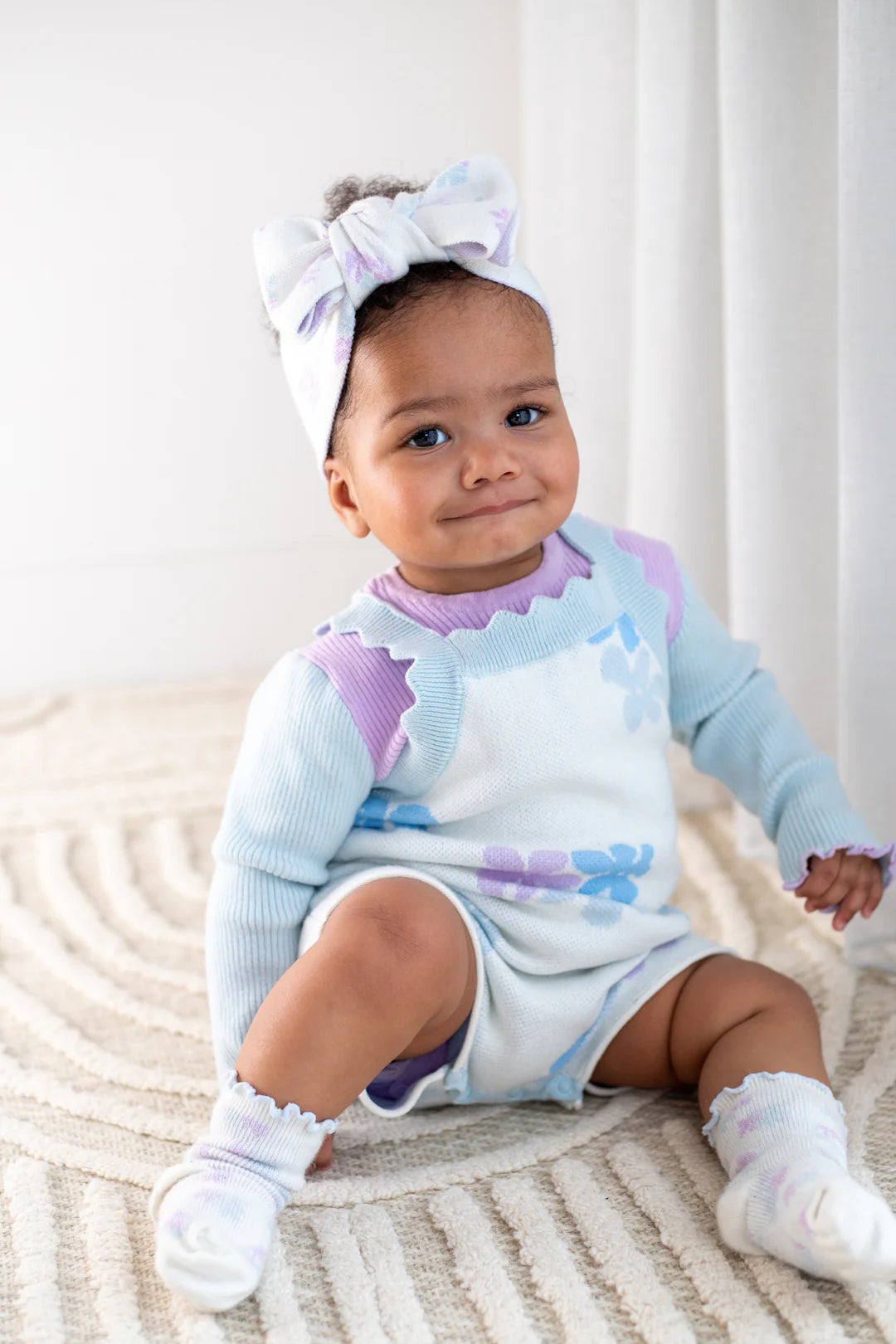 A baby sits on a patterned rug, wearing a pastel-colored ZIGGY LOU Playsuit Alaska with floral designs. The 100% cotton outfit includes a matching headband with a large bow and cozy socks. The baby looks at the camera with a slight smile, and soft, curly hair peeks out from under the headband.