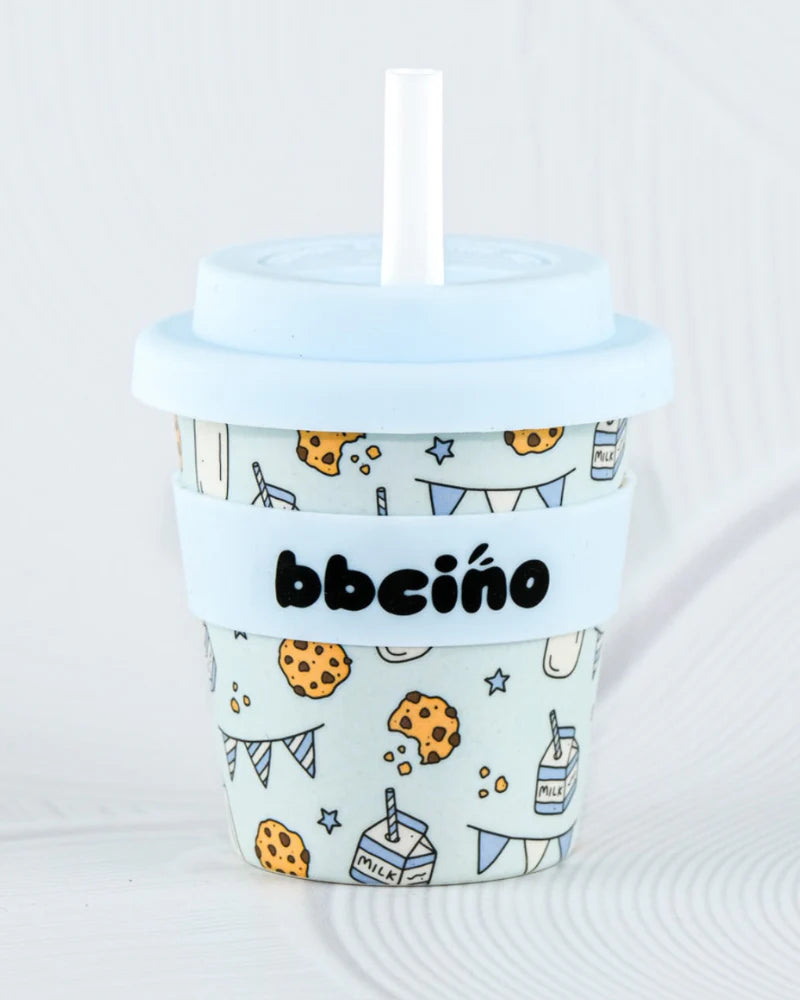 The Reusable Babycino Cup 120ml Tough Cookie in Blue from BBCINO is a small, light blue bamboo fibre cup featuring a playful design of milk cartons, cookies, and party flags. It comes with a lid and a reusable silicone straw, and the text "BBCINO" is elegantly displayed on a matching band around its middle, making it a charming and sustainable choice for babycino cups.