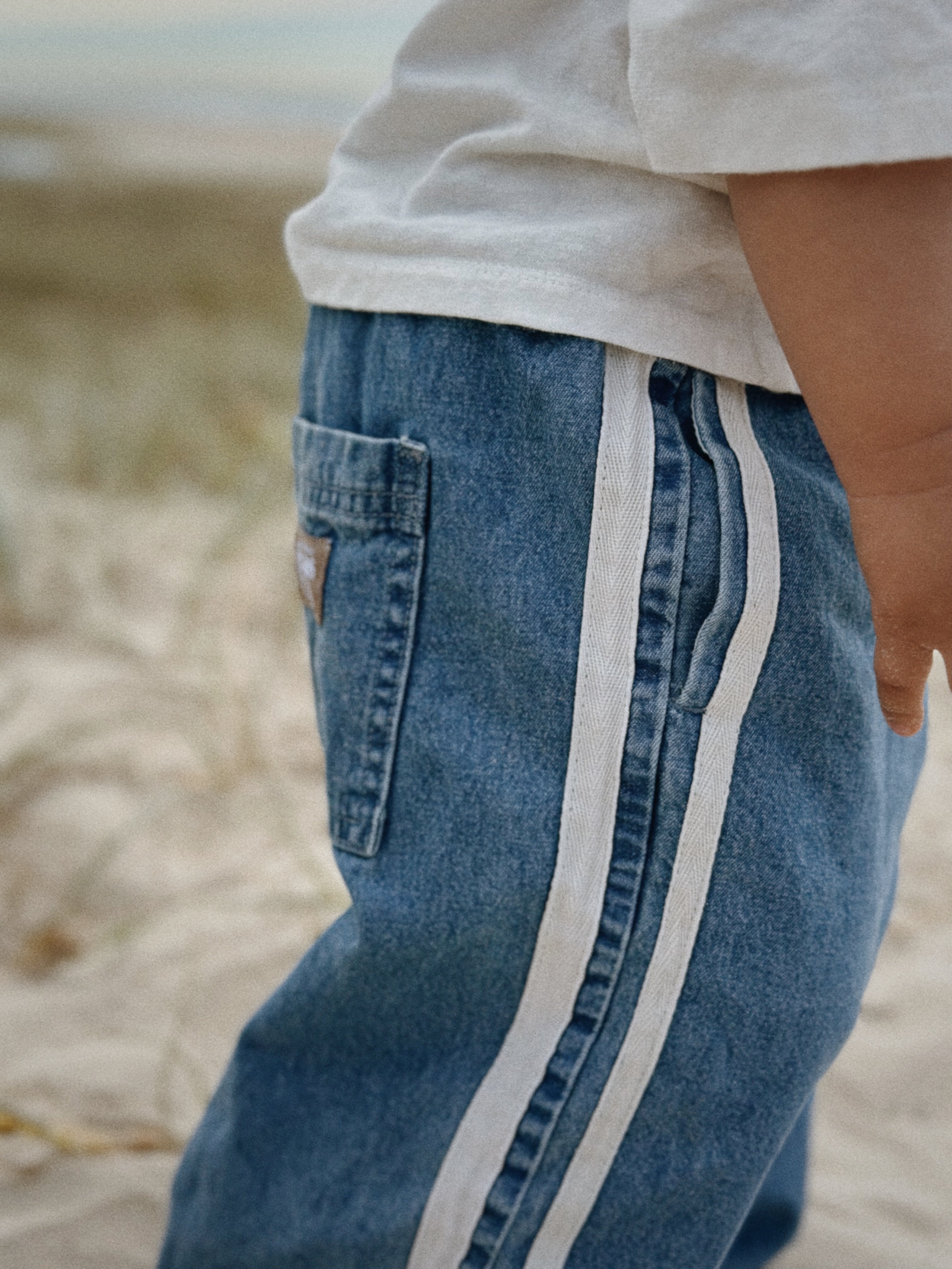 A close-up of a child standing in the sand. The child wears vintage-inspired Tricky Track Pant by TWIN COLLECTIVE, featuring white stripes down the sides, paired with a white T-shirt. Only the lower part of the child's torso and one arm are visible. The background shows a blurred, sandy area, possibly at a beach.