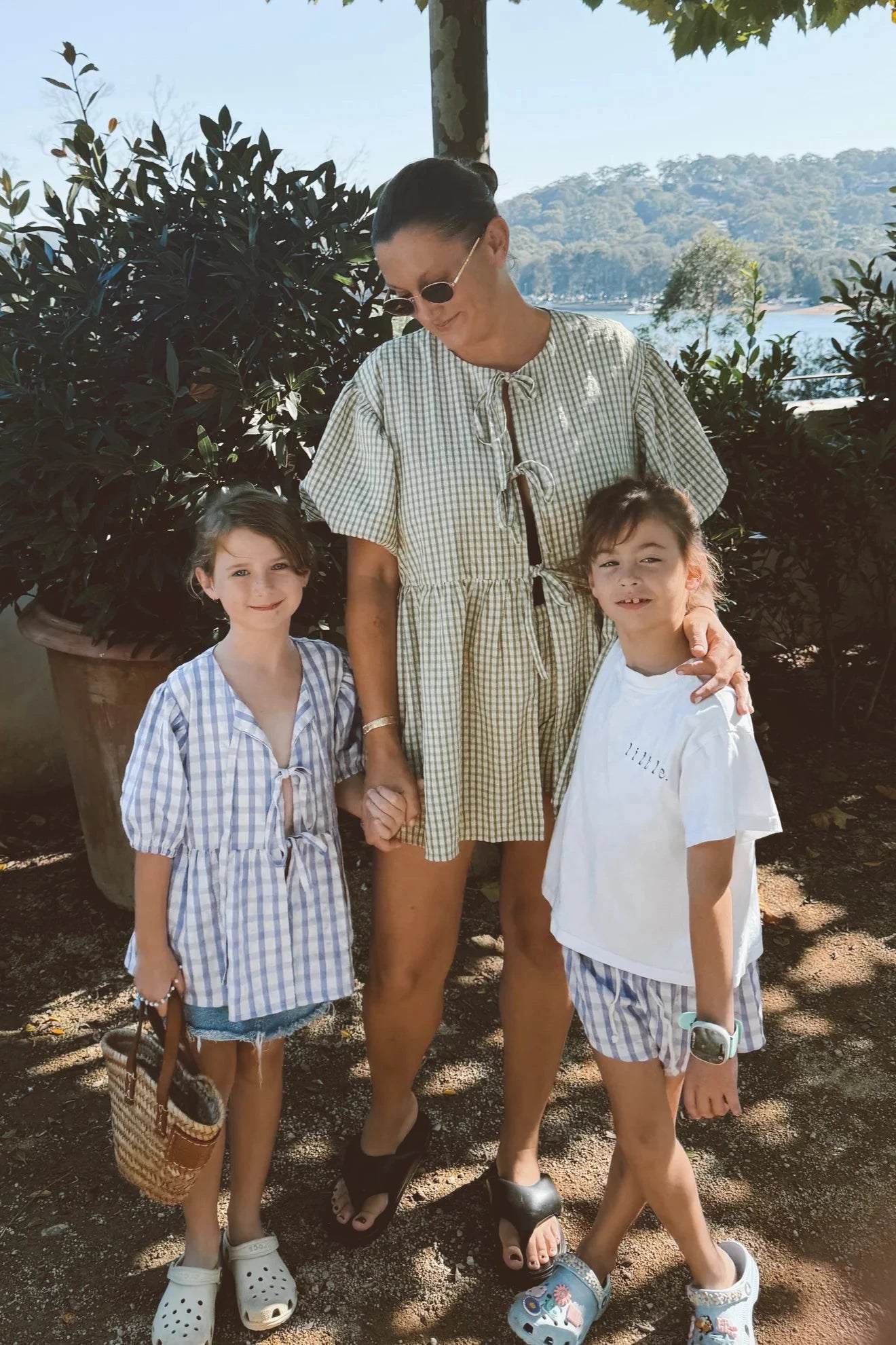 A woman wearing sunglasses stands with her arms around two children in a sunny outdoor setting. One of the children is dressed in a Mama Cami Blouse Olive Gingham from LITTLE THE LABEL, holding a small basket. Trees and a body of water are visible in the background, creating a perfect summer day scene.