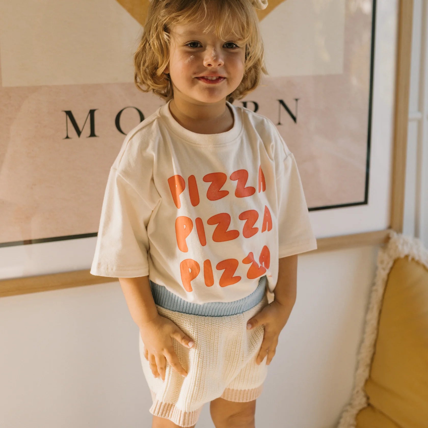 A young, blonde child wears the GOLDEN CHILDREN Pizza Mid Sleeve Tee Eggnog, crafted from soft stretch fabric with "PIZZA PIZZA PIZZA" in red. Light shorts complete the indoor outfit, with a modern framed picture partly visible in the background.