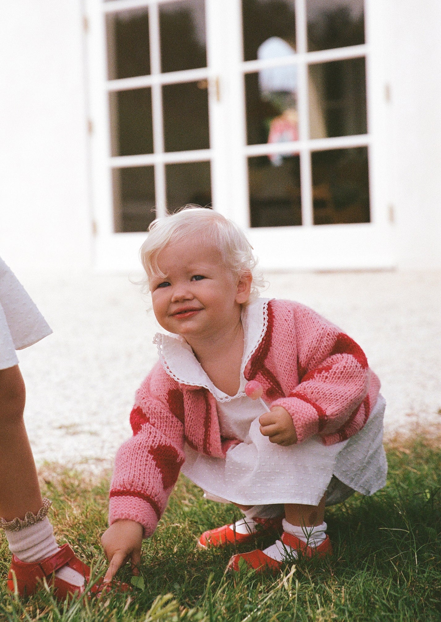A young child with curly blonde hair, wearing THE WHOLESOME STORE's Lover Knit Cardigan Mini in pink, a white dress, and red shoes, squats smiling on green grass. A glass door with a reflection is visible in the background.