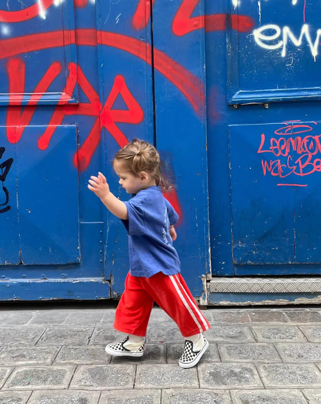 A toddler wearing a Ziggy Zaza Love Your Mother Tee in lapis blue, paired with red pants and checkered shoes, energetically walks down the sidewalk. The backdrop displays a blue wall with vivid red graffiti.