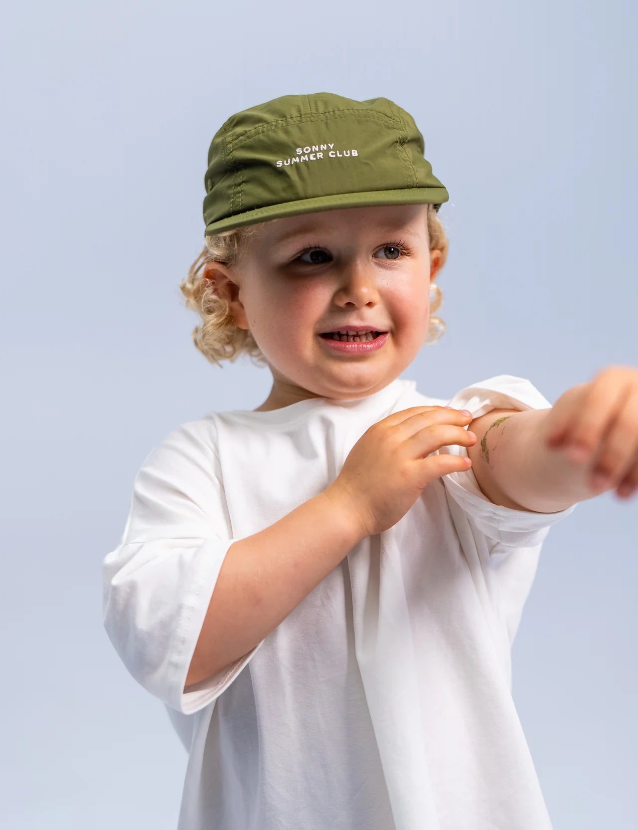 A young child with curly hair wears the Summer Club Cap in Khaki from SONNY LABEL, featuring an adjustable velcro strap, along with a white T-shirt. They smile gently while playfully pointing their forearm forward, set against a light gray background.