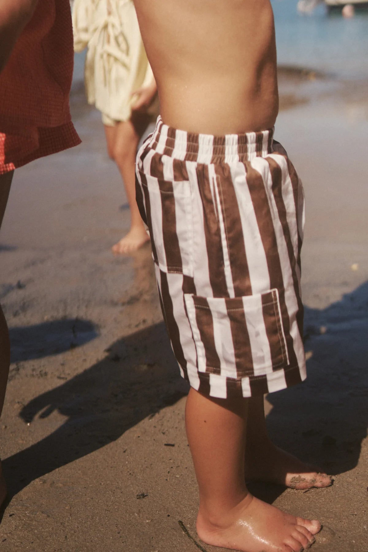 A child in LITTLE THE LABEL's Tei Shorts Brown/White Stripe stands on a sandy beach, feet slightly wet at the water's edge, while others enjoy the sunny day by the ocean in the background.