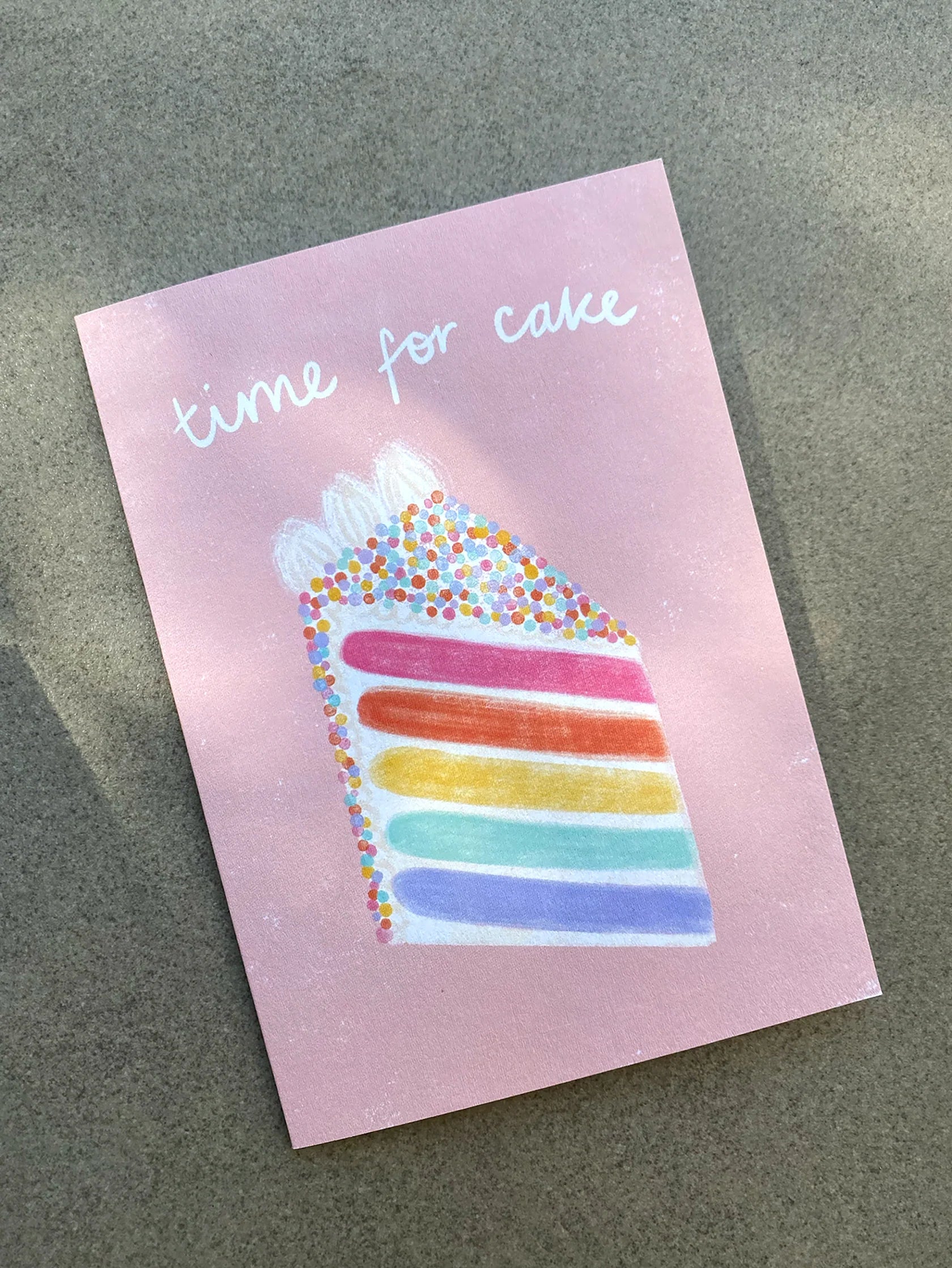 The Birthday Greeting Card "Time For Cake" from LAUREN SISSONS STUDIO features a pastel pink design with an illustration of a slice of rainbow sprinkle cake topped with white icing and colorful sprinkles. The playful white font above the cake reads, "time for cake." Crafted from recycled paper, this card is placed on a grayish surface.