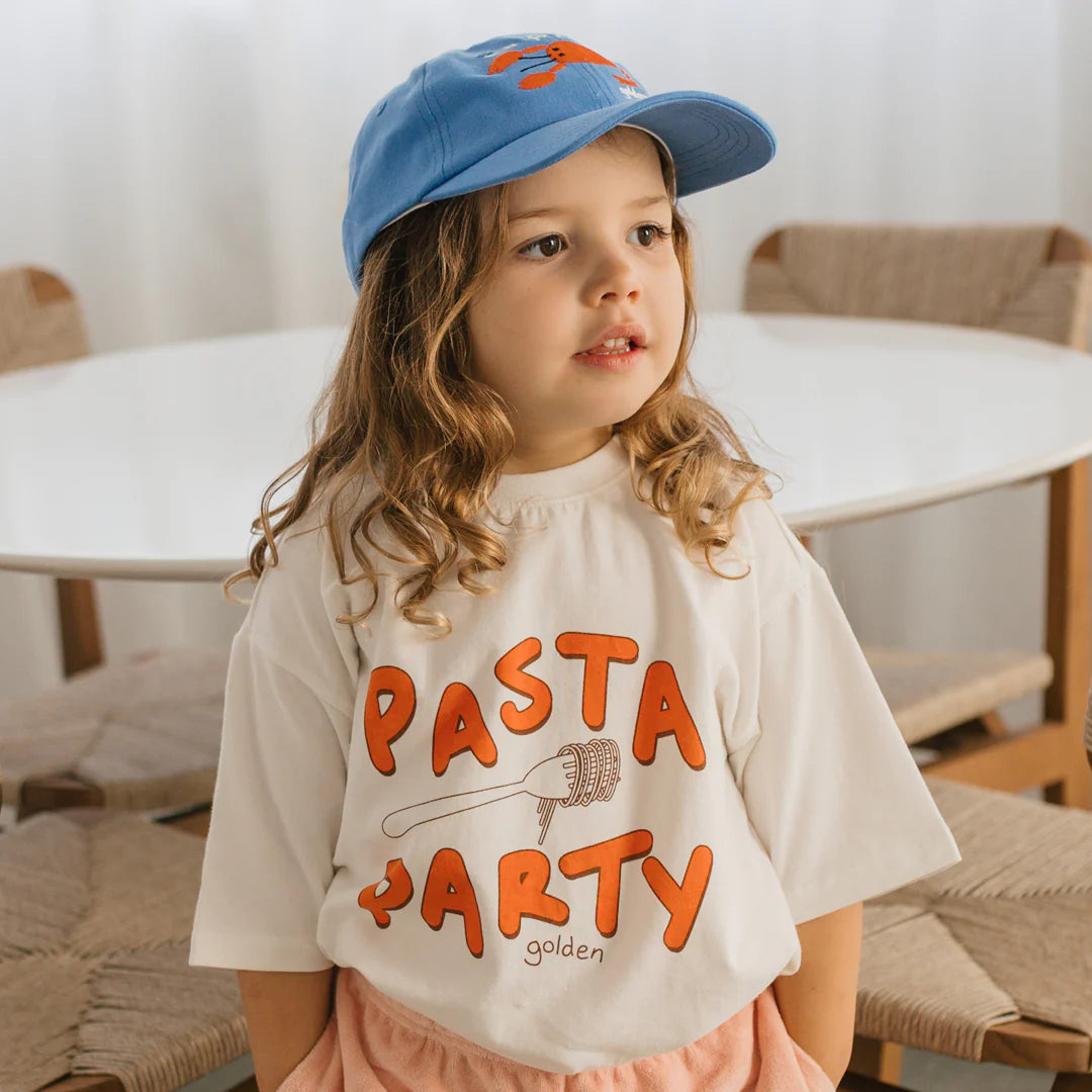 A young child with curly brown hair wears the Lobster Cap in Deep Sea Blue by GOLDEN CHILDREN and a white t-shirt with "Pasta Party" written on it, standing in front of a round white table. The room features neutral-toned chairs and a gentle, soft light ambiance.