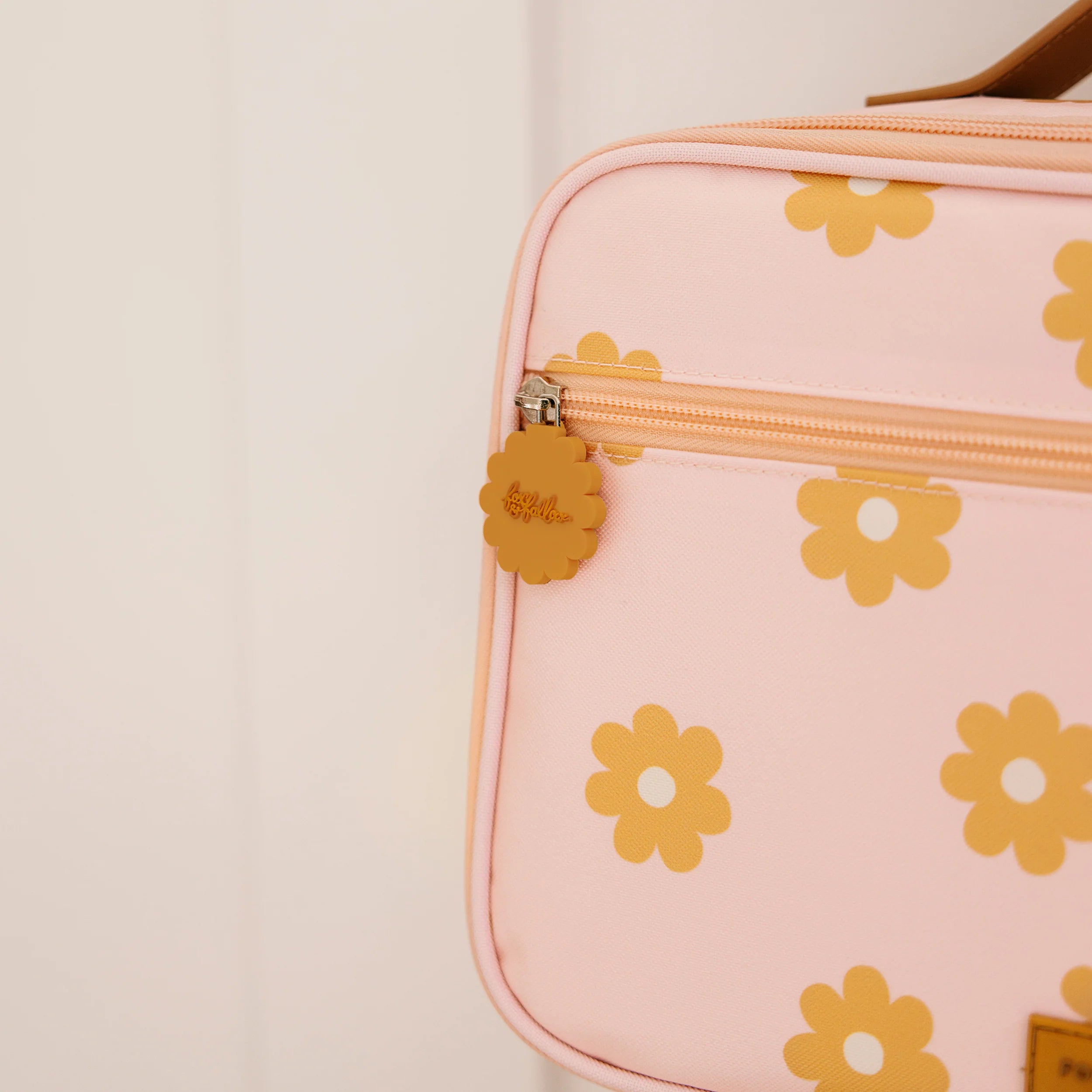 Close-up of a pale pink FOX & FALLOW Daisy Chain Lunch Bag with a front zipper pocket. The bag showcases a pattern of yellow flowers and includes a flower-shaped zipper pull inscribed with "hello simple." The background is off-white.