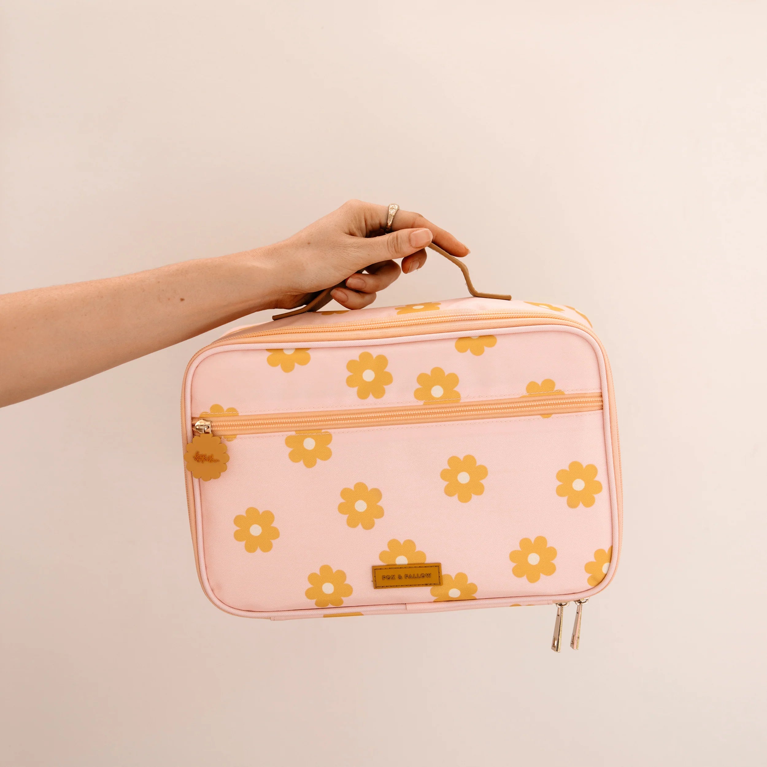 A hand is holding the Daisy Chain Lunch Bag by FOX & FALLOW, featuring a charming pink design adorned with yellow flower patterns. The bag includes a front zipper and a small brown leather label. The plain, light-colored background enhances its appeal, making it as delightful as other insulated lunch bags that are also easy to clean.