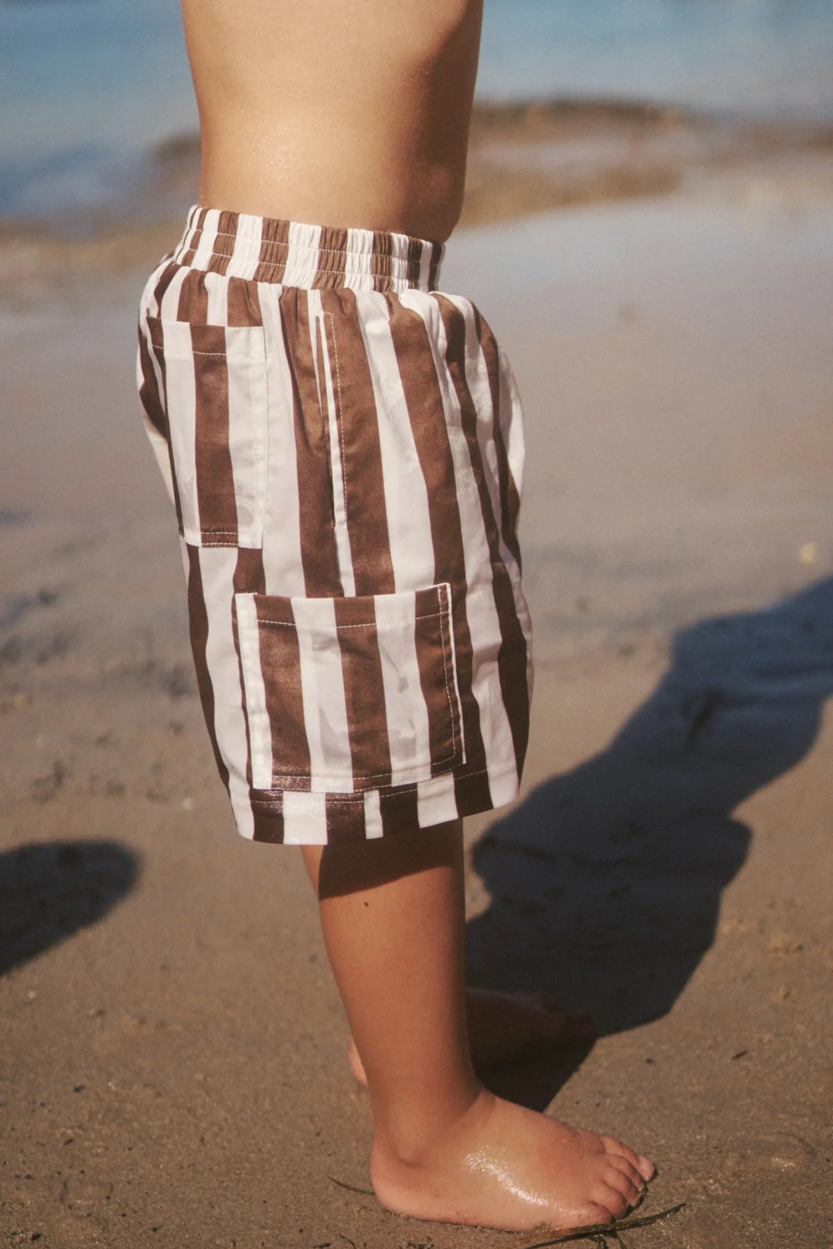 A child stands barefoot on a sandy beach, near the water's edge, wearing the Tei Shorts in Brown/White Stripe by LITTLE THE LABEL. Shadows are cast on the sand.