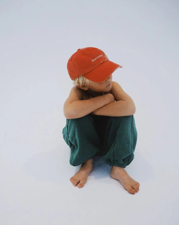 A blonde child, sitting cross-legged on a plain white floor, wears the stylish Ziggy Zaza ~ Roses Are Red Cap Washed Red and green pants. The cap's adjustable brass clasp stands out as they gaze to the side, arms crossed over knees.