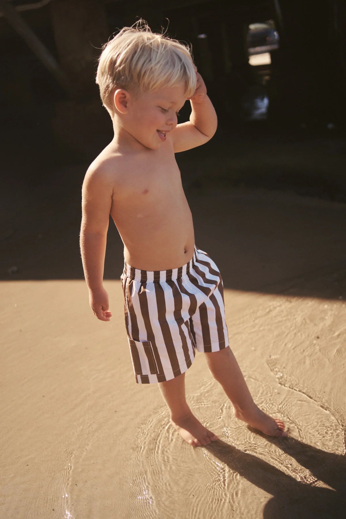 A young blonde boy stands playfully on a sandy beach wearing LITTLE THE LABEL's Tei Shorts in Brown/White Stripe. He touches his head with one hand, looking down as gentle waves lap at his feet under the warm sunlight.