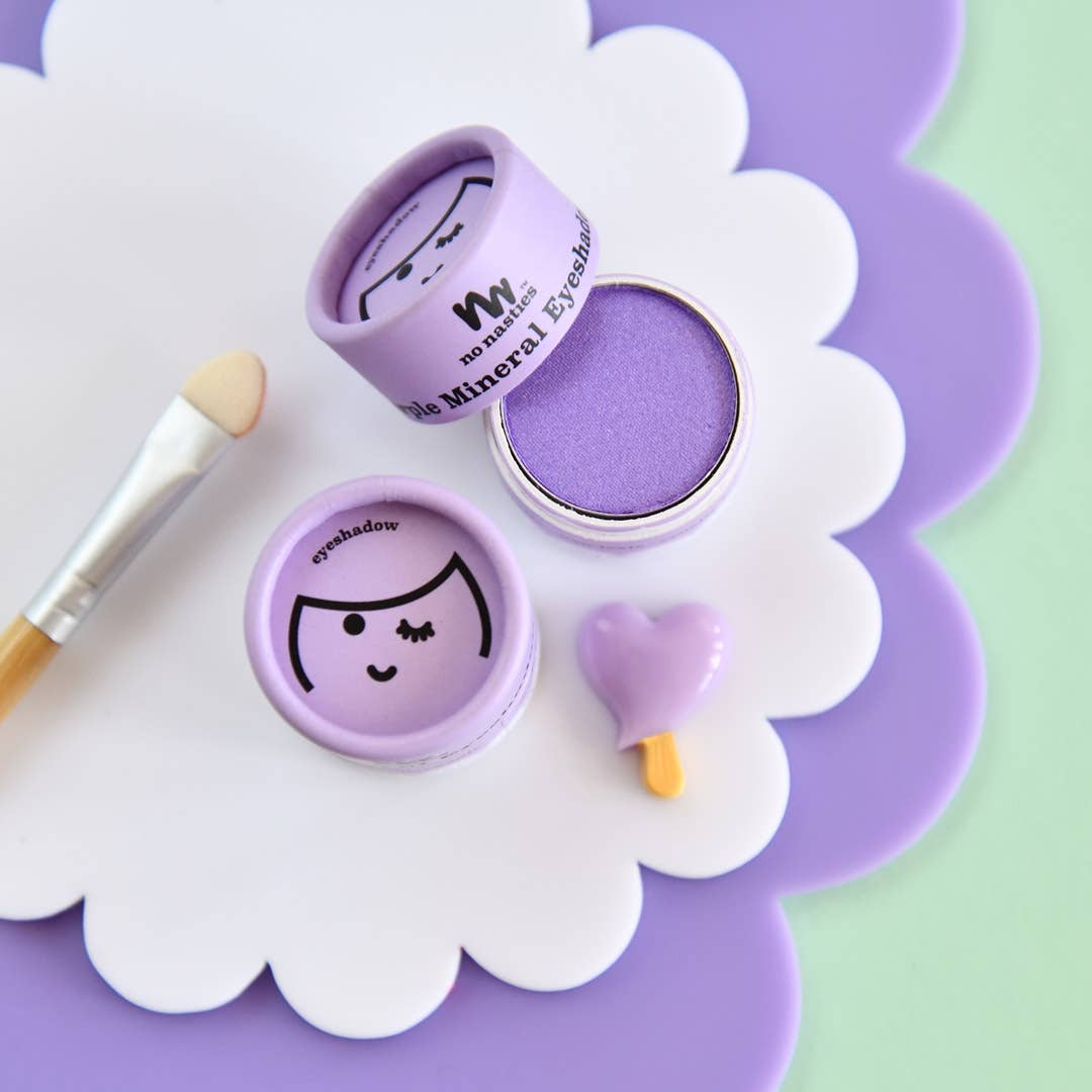 A pastel-themed flat lay features an open container of Kids Eco Natural Pressed Eyeshadow & Blush by no nasties kids with a smiling face design, a heart-shaped makeup brush cleaner, and an eyeshadow brush. Made with natural ingredients and a hypoallergenic formula, the background consists of pastel green and purple surfaces with white scalloped detailing.