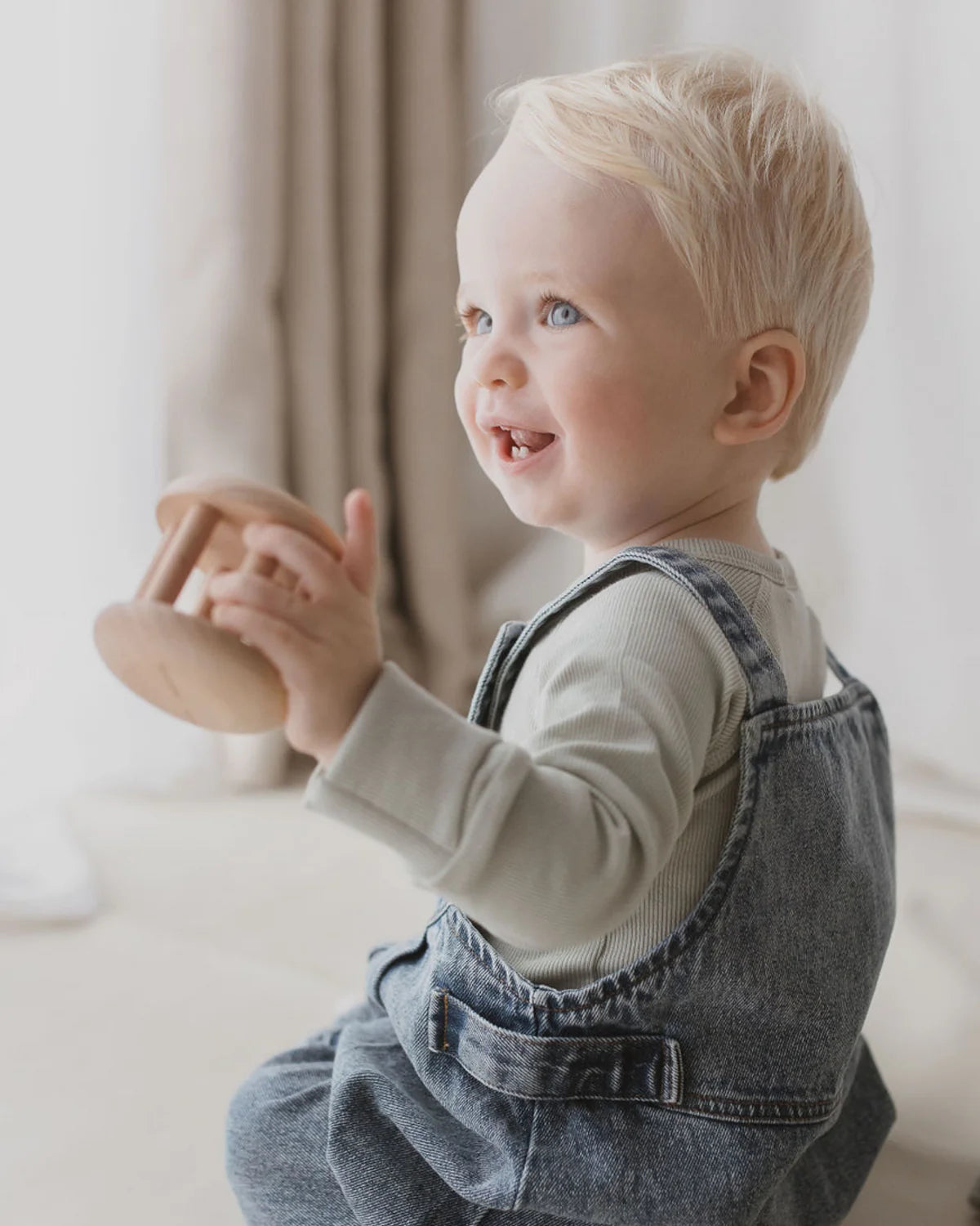 A smiling toddler with light blonde hair and blue eyes is holding a wooden toy. The child, dressed in a SUSUKOSHI light-colored long-sleeve shirt and Denim Overalls Sky Blue with adjustable shoulder straps, sits indoors against a softly lit background with beige curtains.