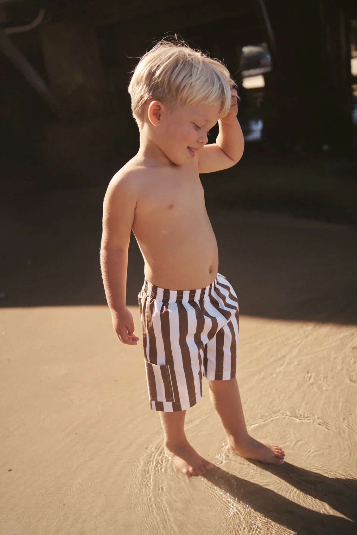 A young child stands on a sandy beach wearing Tei Shorts Brown/White Stripe by LITTLE THE LABEL. The child gently touches their head while gazing downward, with sunlit sand and a shadowy area in the background.