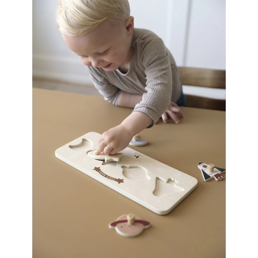 A young child with blonde hair is playing with a FABELAB Grip Puzzle Space Magic at a light brown table. The puzzle pieces include a star, moon, and other shapes. Focused on placing a piece into the correct slot, the child demonstrates fine motor control. The room background is minimal with soft, neutral tones.