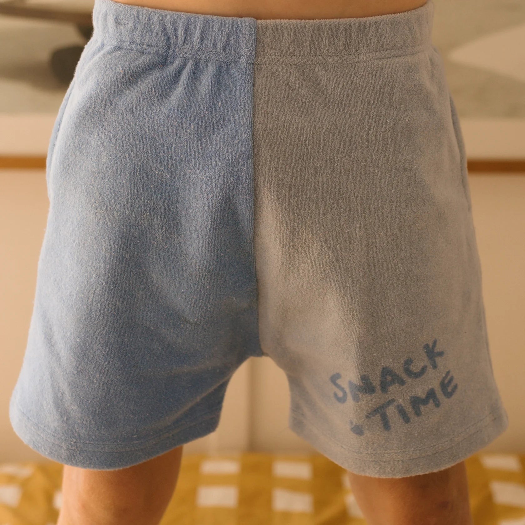 A person wearing GOLDEN CHILDREN Play Lounge Shorts Pool, featuring a two-toned blue design in terry towel cotton. The left side of the shorts is a darker blue, while the right side is a lighter blue with "SNACK TIME" printed on it. In the background, part of a bed with a yellow checkered blanket is visible.