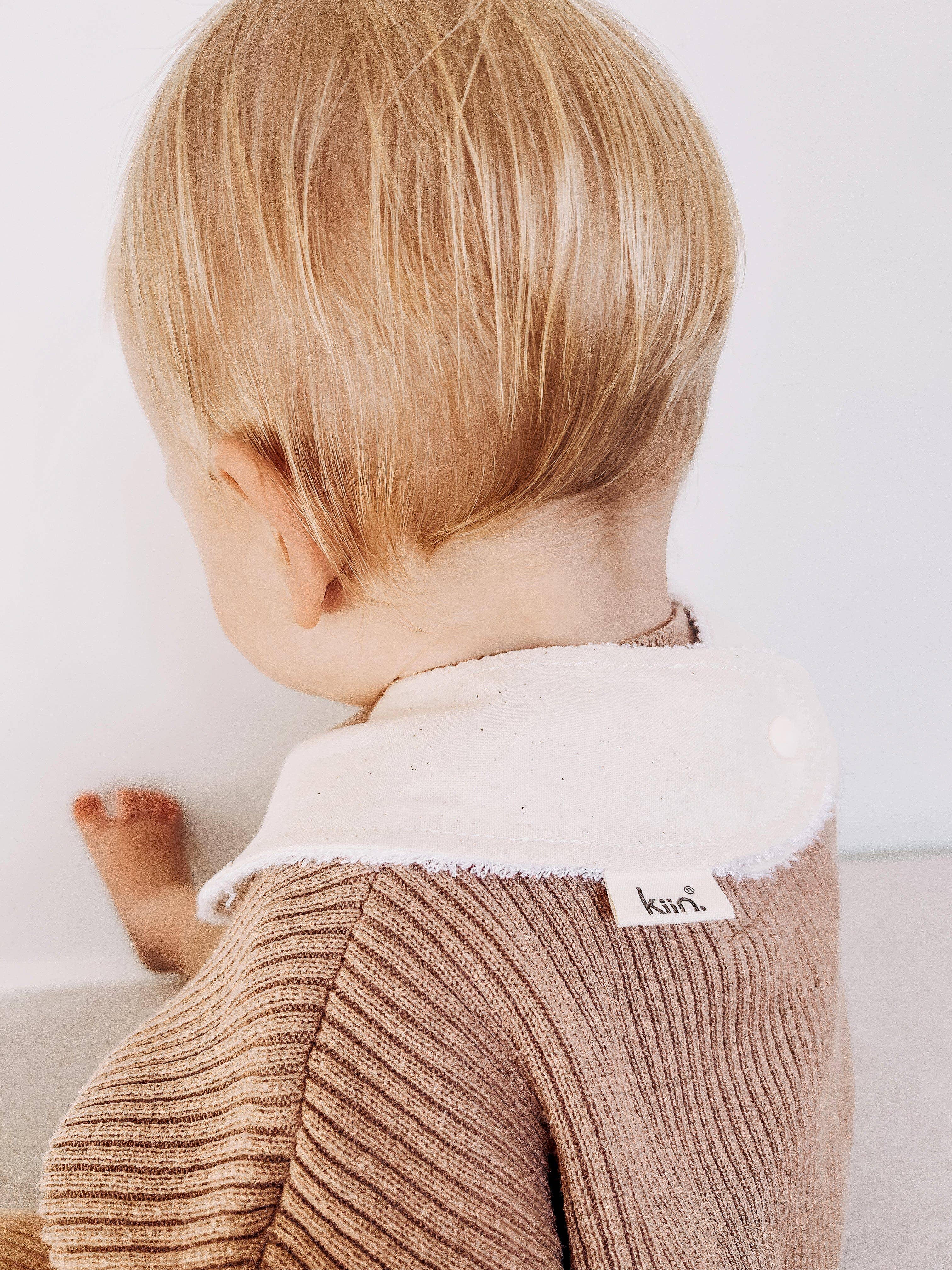 A toddler with light blonde hair wears a brown ribbed sweater with a white collar and KIIN BABY's Dribble Bib Oat. The photo captures the child's head and upper body against a light background.