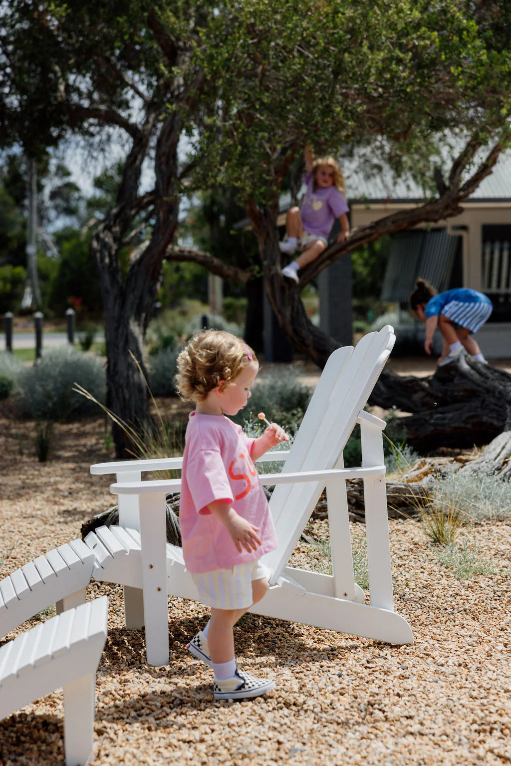 A young child wearing a slightly oversized Sunday Siblings Il Sole Tee in Pink/Red stands near a white wooden chair on gravel. Two other children play nearby, one climbing a tree and another on a large rock, with trees and a building as the backdrop for their carefree play.