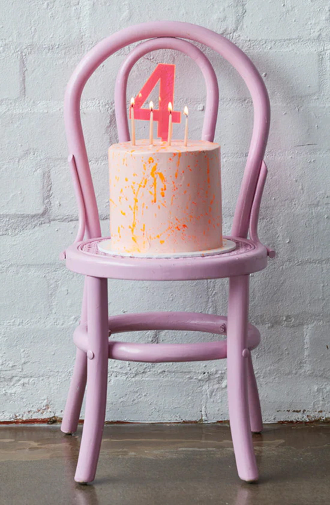 A HELLO KIT CO Large Cake Topper in a Neon Pink colour on top of a pink cake splattered with orange icing on a pink chair against a white brick wall.