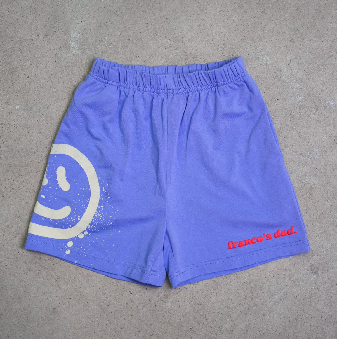 Acid Dye Shorts Blue by FRANCO'S DAD are designed in stretch cotton with a unique purple hue. They feature a white smiley face on the left side and the text "Franco's Dad." in red on the right. These comfortable shorts, styled like an acid dye lounge set, are photographed flat on a textured gray surface.