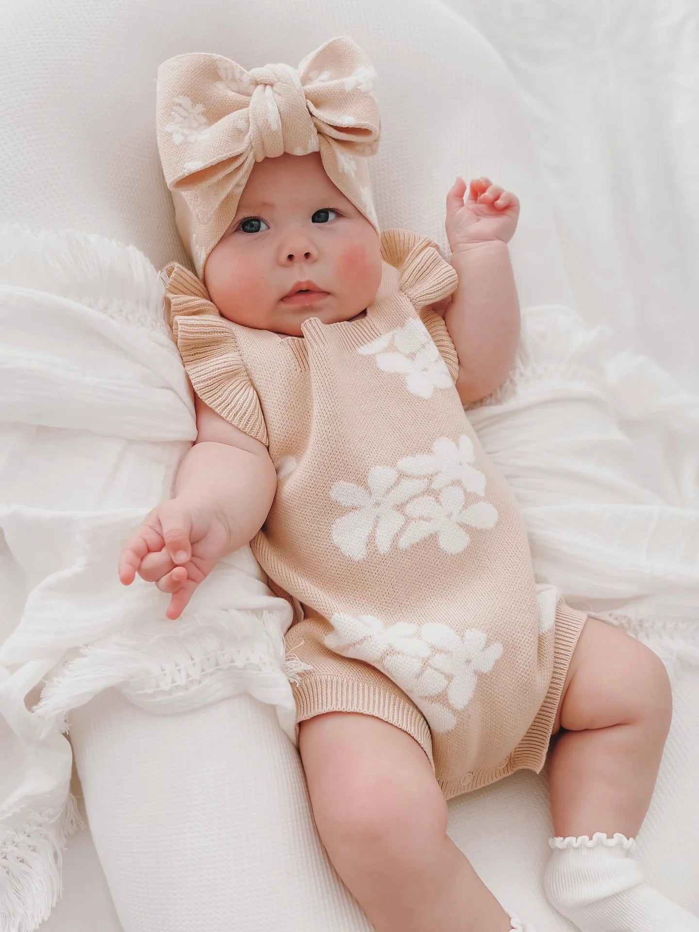 A baby dressed in a light brown ZIGGY LOU Frill Romper Banksy with white floral patterns lies on a white cushion. The matching headband with a large bow completes the adorable look. The baby's eyes are open and an arm is extended upward, set against a background of soft white fabric.