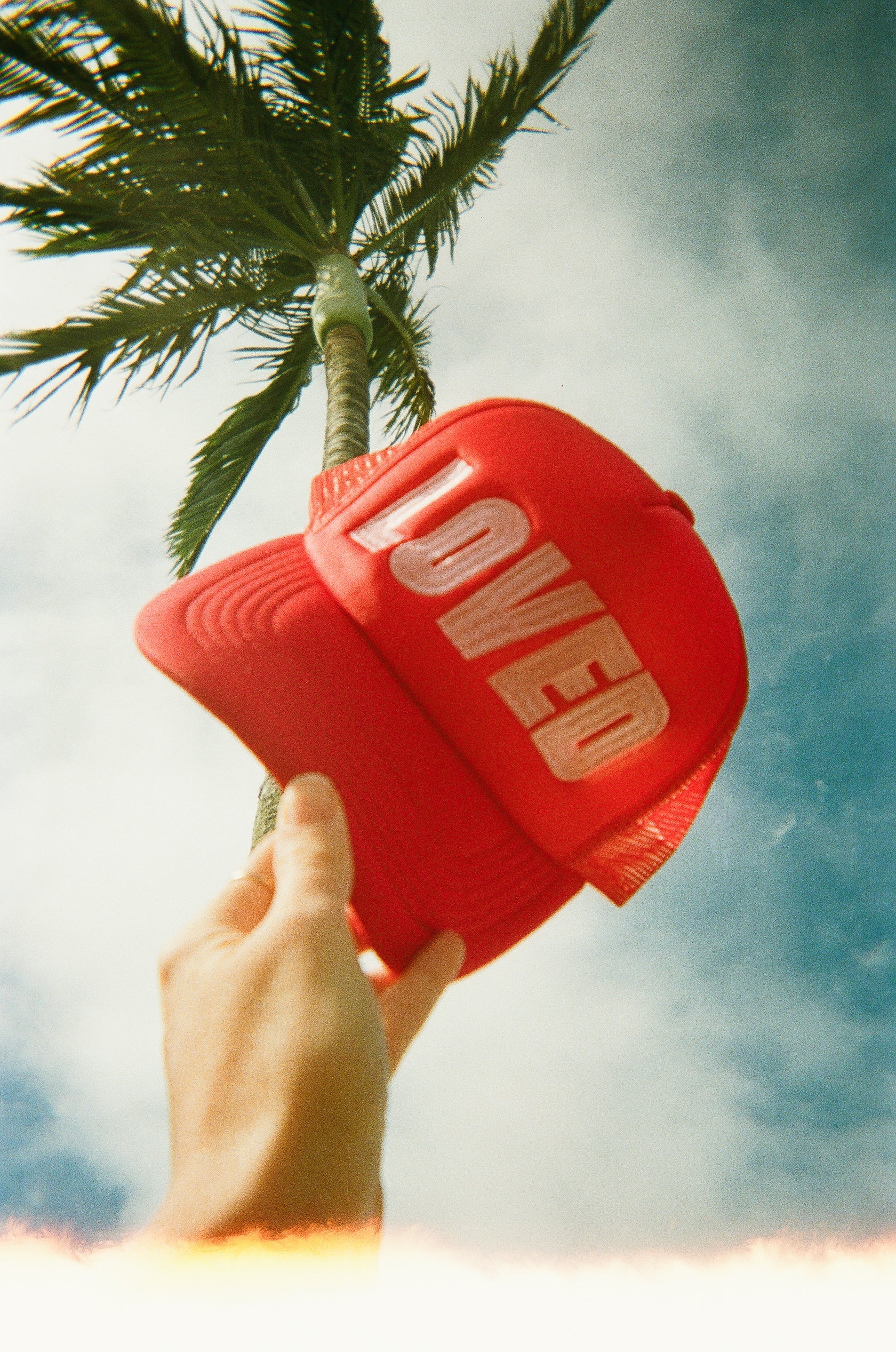 A hand holds up the TINY LOVE CLUB's So Loved Trucker Cap, featuring "LOVED" in bold letters. This sun-safe cap with an adjustable back strap stands out against a partly cloudy sky and a gently swaying tall palm tree.