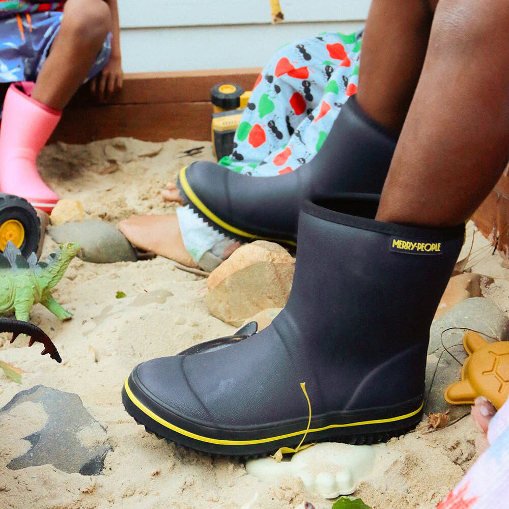 Children wearing Merry People's Andie Kids Gumboot in Midnight Blue play in a sandbox with dinosaurs and toy trucks. One sports black boots with yellow trim, another dons pink vegan boots. Seashells and rocks scattered in the sand enhance their adventure.