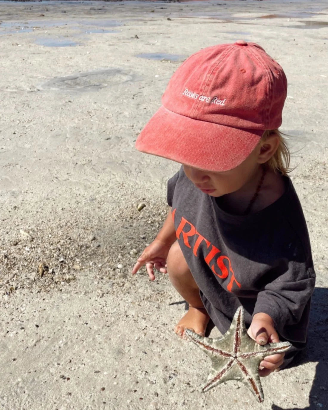 A child in a washed red Ziggy Zaza cap labeled "Roses Are Red" and an oversized dark shirt crouches on a sunlit beach, clutching a starfish. The cotton canvas cap, fastened by an adjustable brass clasp, partly obscures the shimmering shoreline in the background.