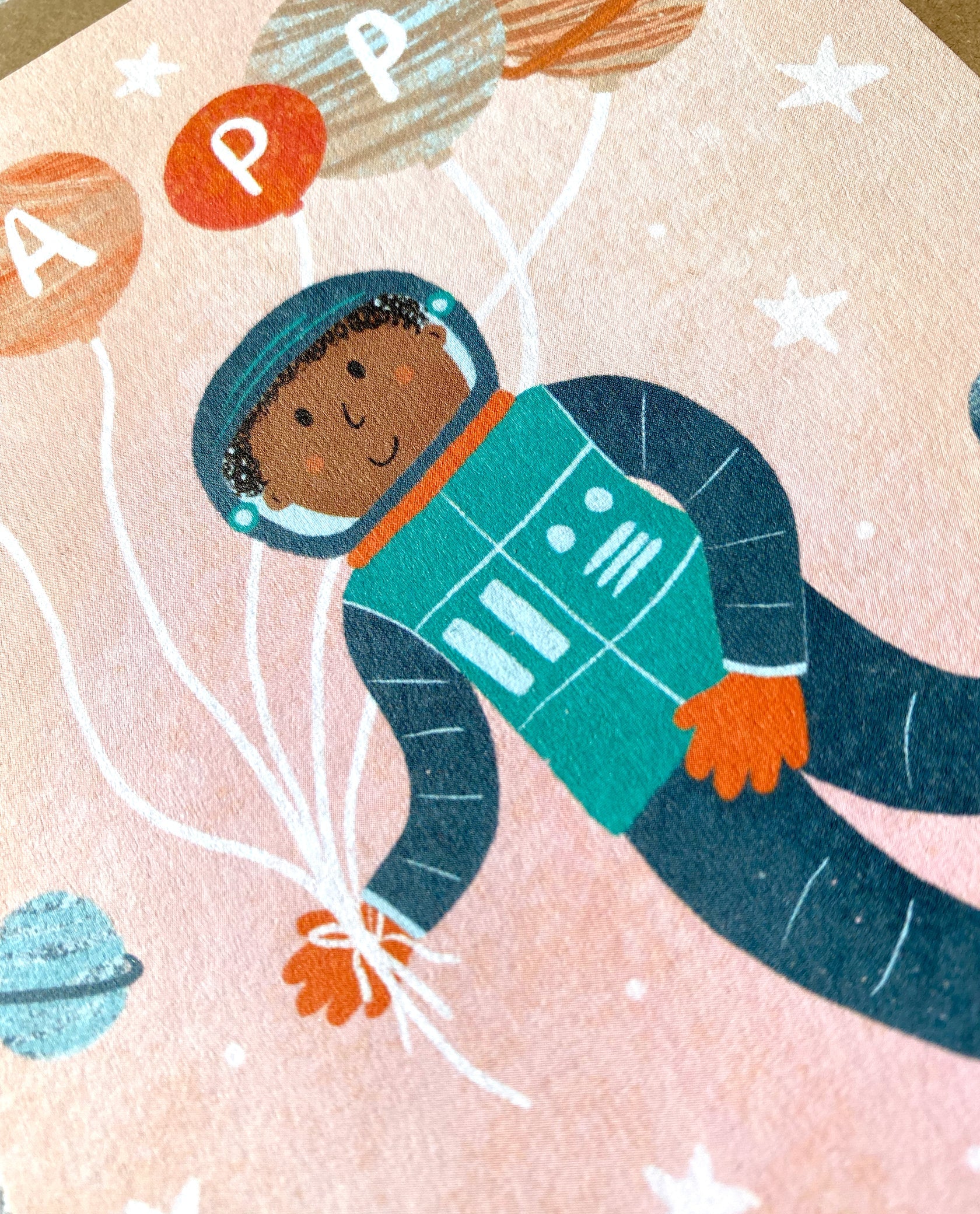 A cheerful astronaut floats in space, surrounded by stars and planets. The astronaut is holding a bunch of balloons with letters on them spelling "HAPPY," wearing a space suit, and smiling against a light peach background—perfect for an outer space themed card for a children's birthday celebration. This delightful scene is beautifully captured on the Birthday Greeting Card “Astronaut” by LAUREN SISSONS STUDIO.