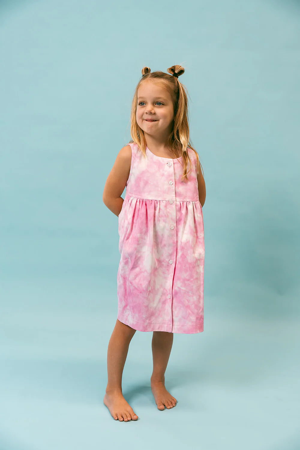 A young girl stands barefoot on a light blue background, wearing the Lollipop Dress from LENN LABEL. Her pink tie-dye dress features a relaxed fit. She has light brown hair styled in two small buns and smiles while looking off to the side with her hands behind her back.