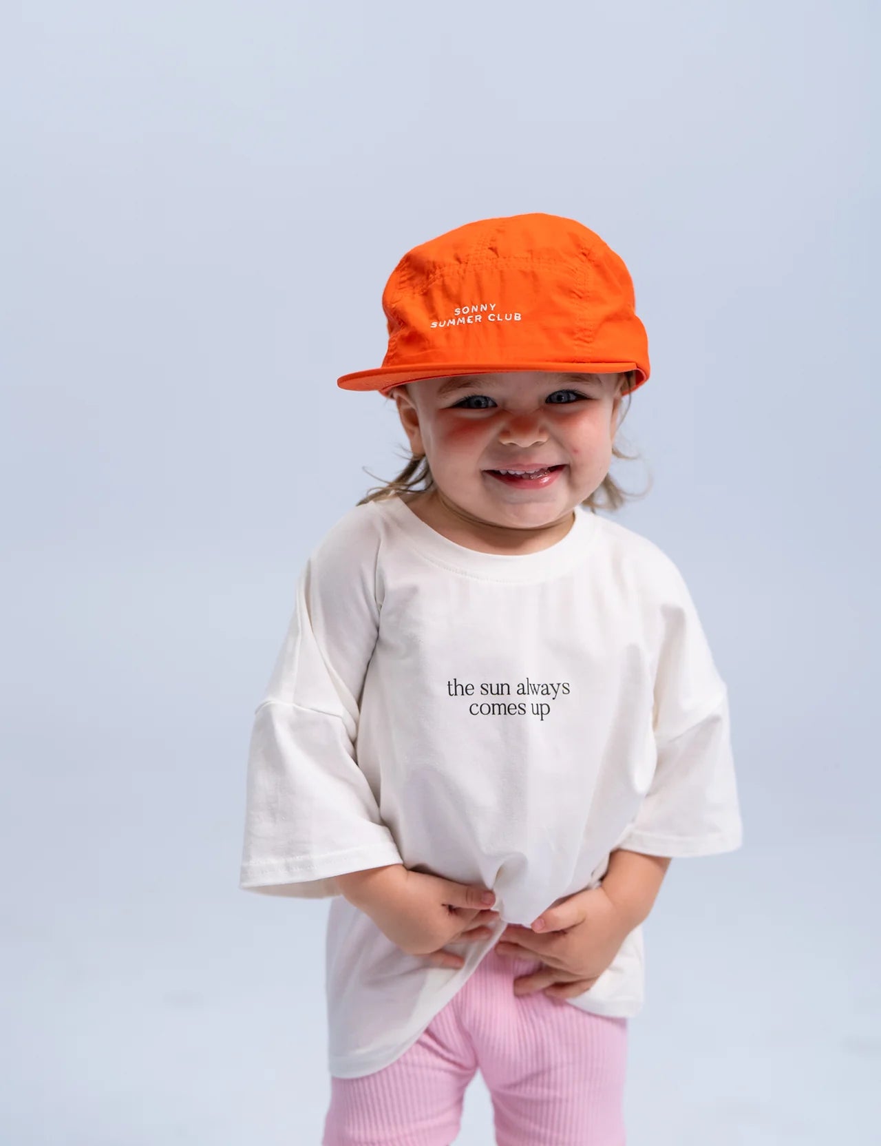 A young child wearing the SONNY LABEL Summer Club Cap in Poppy, featuring an adjustable Velcro strap, and an oversized white t-shirt with the slogan "the sun always comes up," stands against a light gray background. The child is smiling and wearing light pink pants.