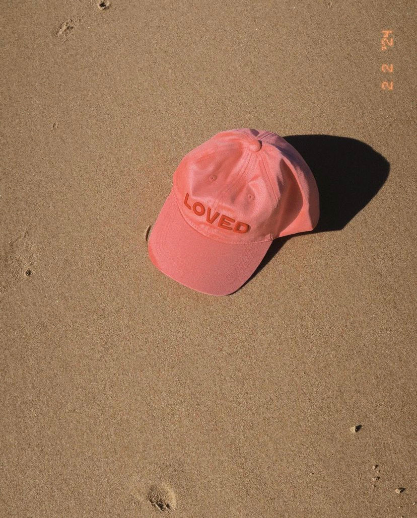 A TINY LOVE CLUB Loved Hat - Pink, featuring the word "LOVED" embroidered on the front, lies on a sandy surface under direct sunlight. The shadow of the cap is visible on the sand, with a few small impressions scattered nearby. Designed in a classic dad style with an adjustable back strap perfect for little heads.