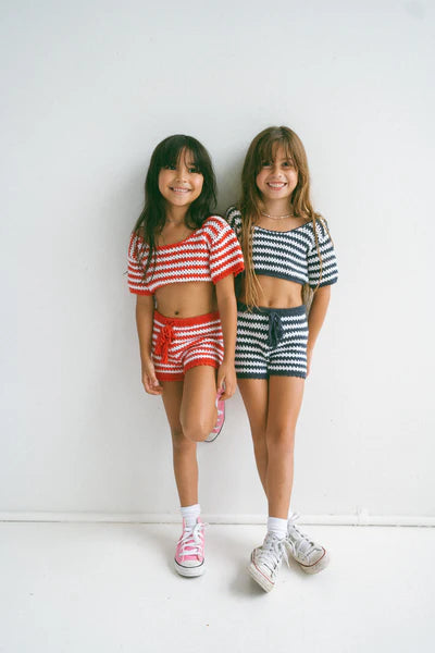 Two children stand against a white background wearing matching NOTHING BUT AMOR Crochet Stripes Sets; one red/white, the other navy/white. Both have long hair and are smiling, their sneakers and soft cotton socks capturing a breezy Bali vibe.