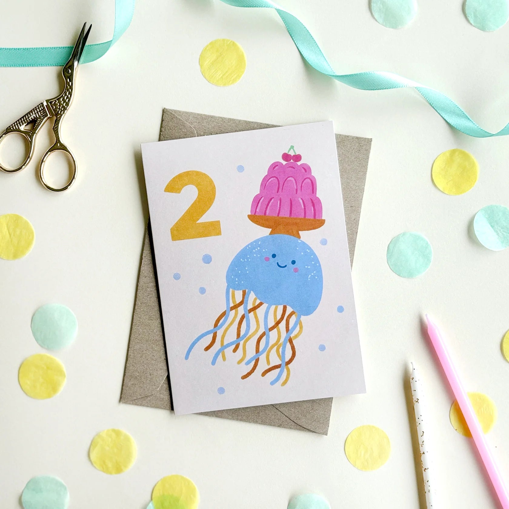 The 2nd Birthday Age Milestone Card featuring an illustrated jellyfish balancing a plate of jelly and two cherries alongside a bold hand-lettered number 2 on a white background. There is blue and yellow paper cut outs around the card with a pair of gold scissors and pencils.