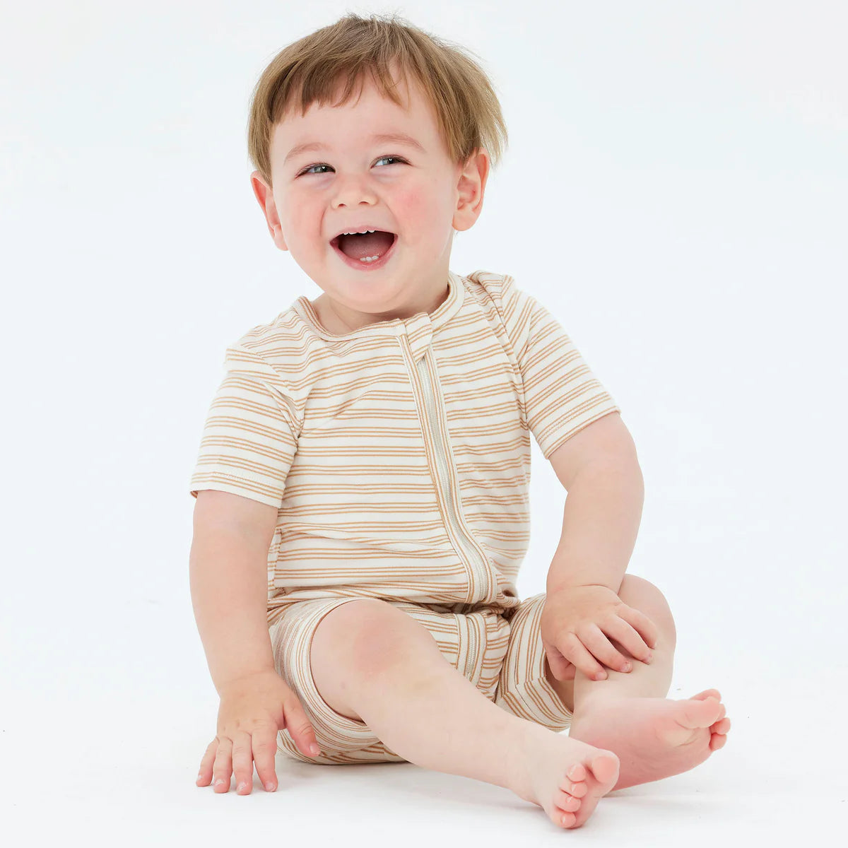 A joyful toddler wearing the KYND BABY Day or Night Short Sleeve Onesie Neutral sits on the floor, laughing happily against a plain white background. Made from sustainable viscose fabric, this beige and white striped outfit provides both comfort and eco-friendly style.