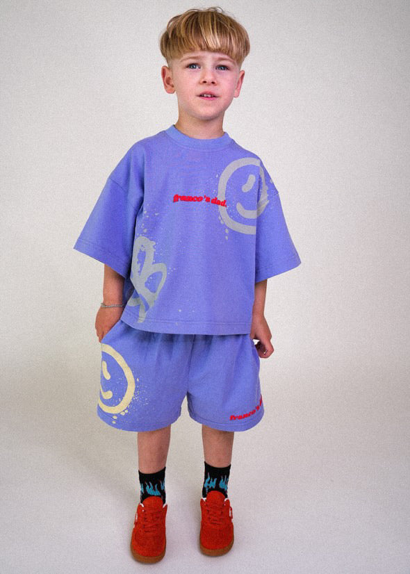 A young boy stands against a plain background, sporting a loose-fitting lounge set in stretch cotton, featuring the FRANCO'S DAD Acid Dye Shorts in blue and a purple shirt with graphic designs like smiley faces. He completes the look with red sneakers and dark socks showcasing flame patterns.