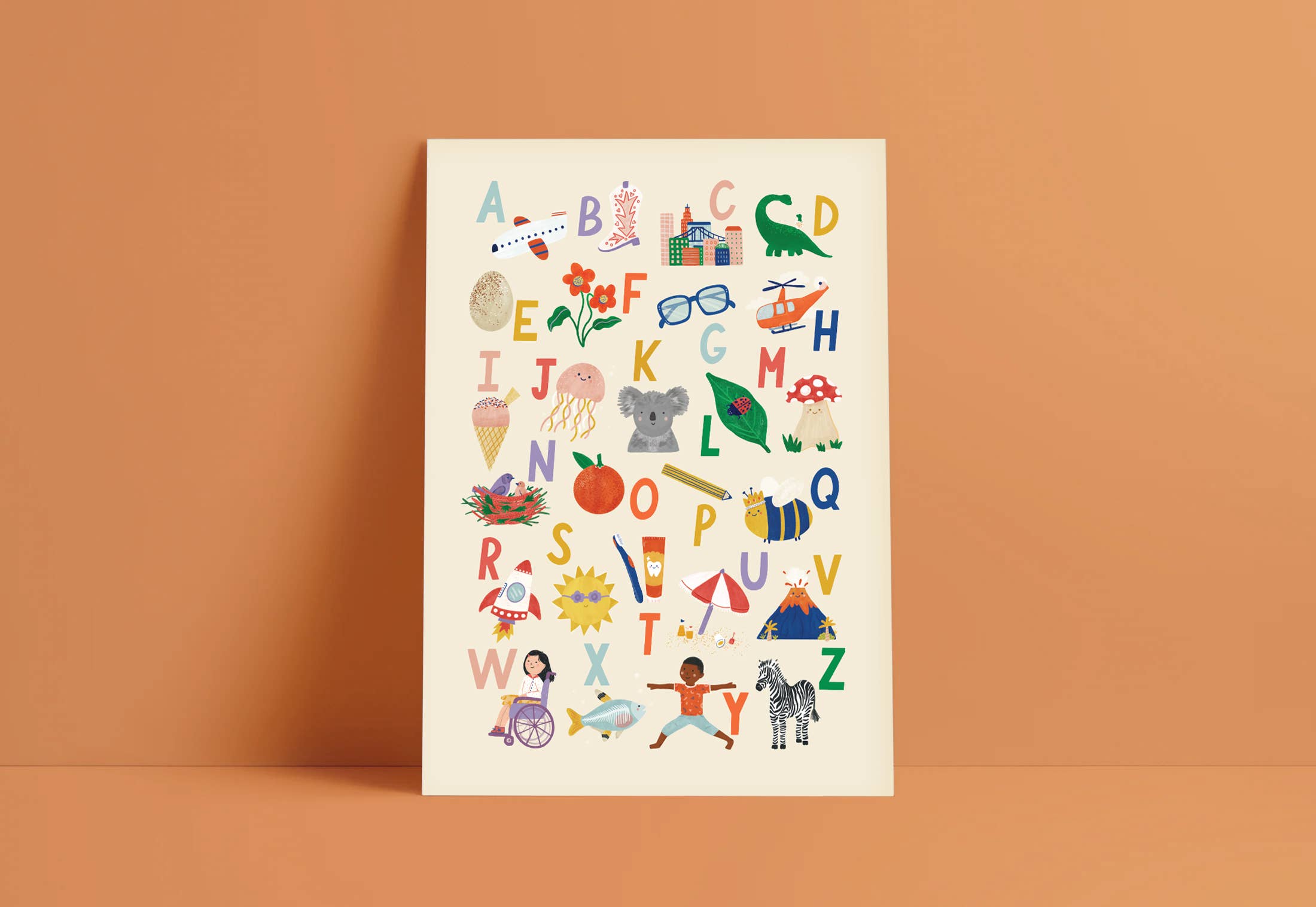 The Illustrated Alphabet Wall Art Print by LAUREN SISSONS STUDIO features a vibrant A-Z alphabet design set against a peach background. Each letter from A to Z is adorned with an adorable illustration, such as an apple for A, a koala for K, and a zebra for Z. Ideal for nursery decor, the poster is displayed propped up and leaning slightly against the wall.