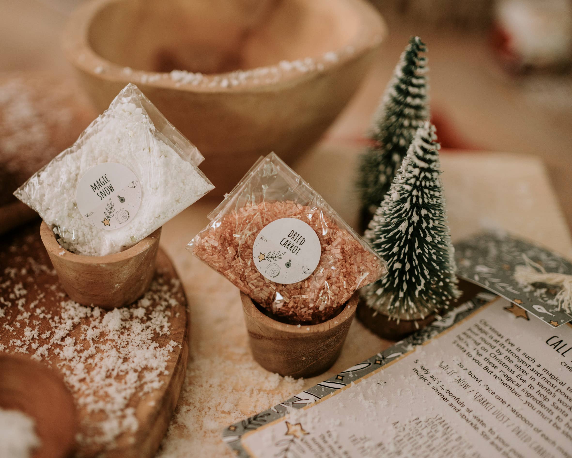 In a cozy scene, two small wooden bowls from THE LITTLE POTION CO's Xmas Eve Potion Pouch to Call the Reindeer are labeled "Magic Snow" and "Dried Caramel." Miniature Christmas trees lend a festive touch as a parchment covered in snow-like powder, containing holiday recipes, lies nearby.