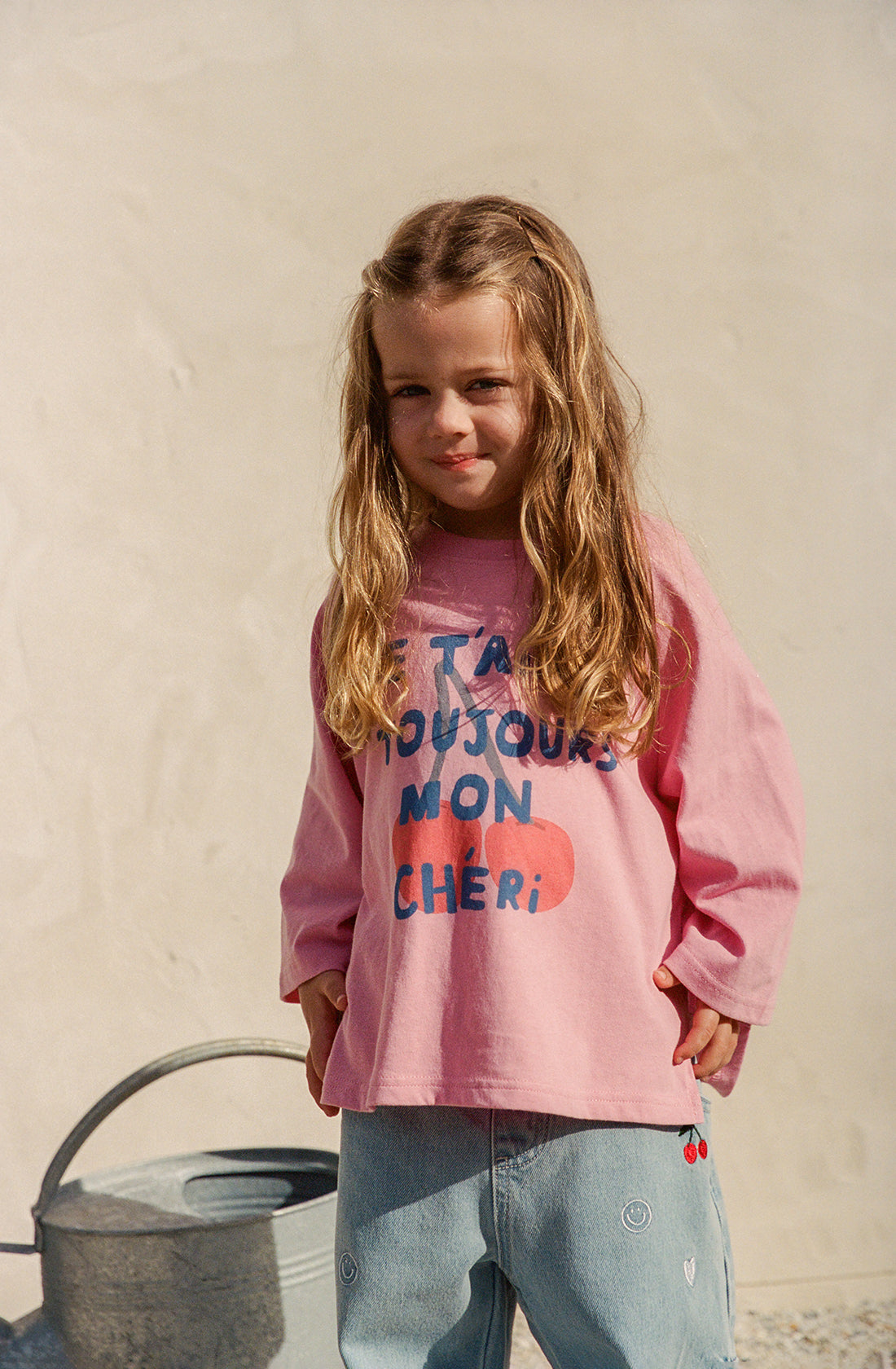 A young child with long, wavy hair smiles softly while standing outdoors. The child is wearing THE WHOLESOME STORE's Je T’aime Long Sleeve Tee Mini in an oversized fit, which reads "TA TOUJOURS MON CHÉRI." They are also dressed in light blue jeans with small embroidered details. Made from 100% GOTS certified cotton, a watering can is visible in the background.