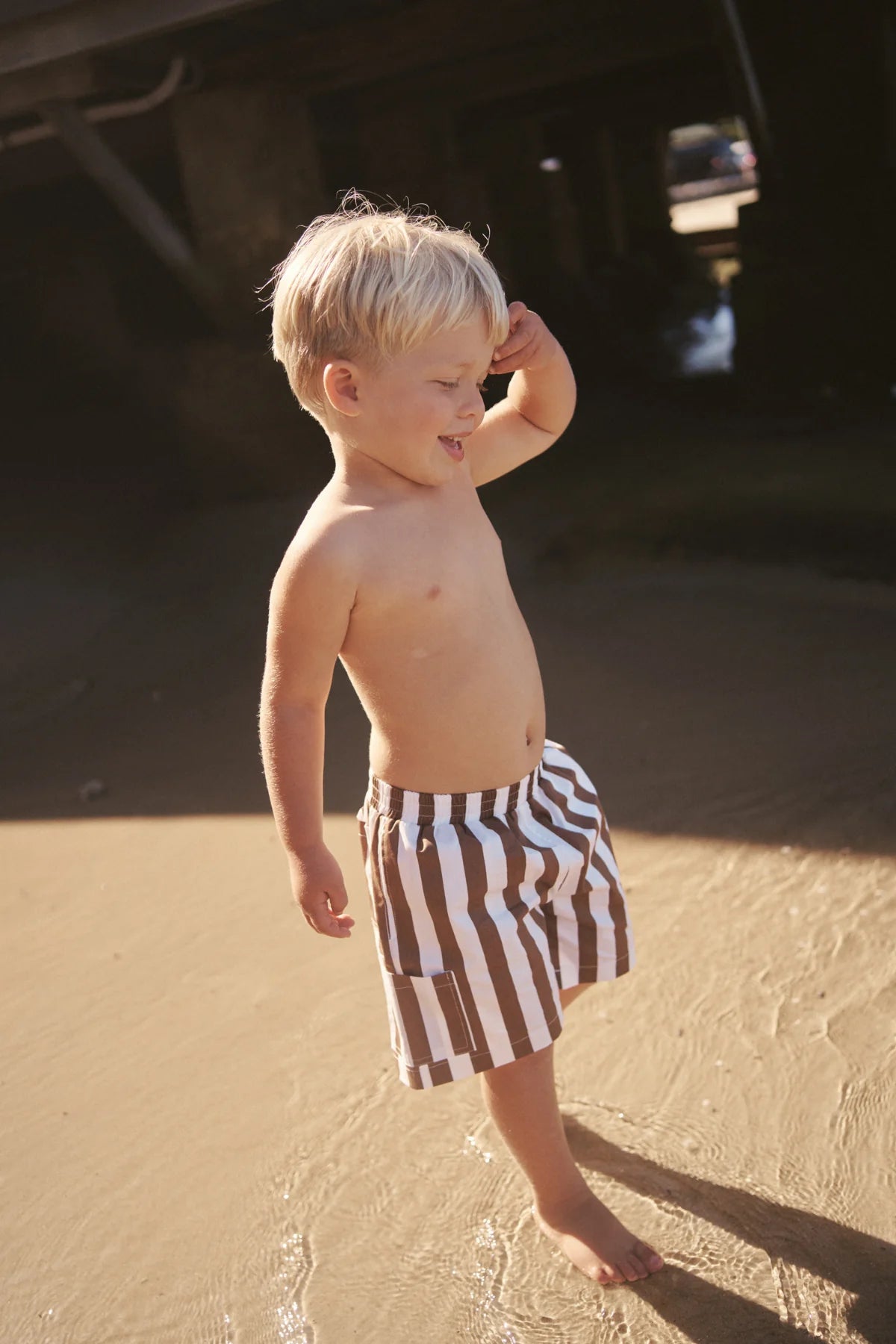 A young blond-haired child stands on a sandy beach, smiling and enjoying the sunny weather. They wear LITTLE THE LABEL's Tei Shorts in Brown/White Stripe as water touches their feet and shadows from a nearby structure form in the background.