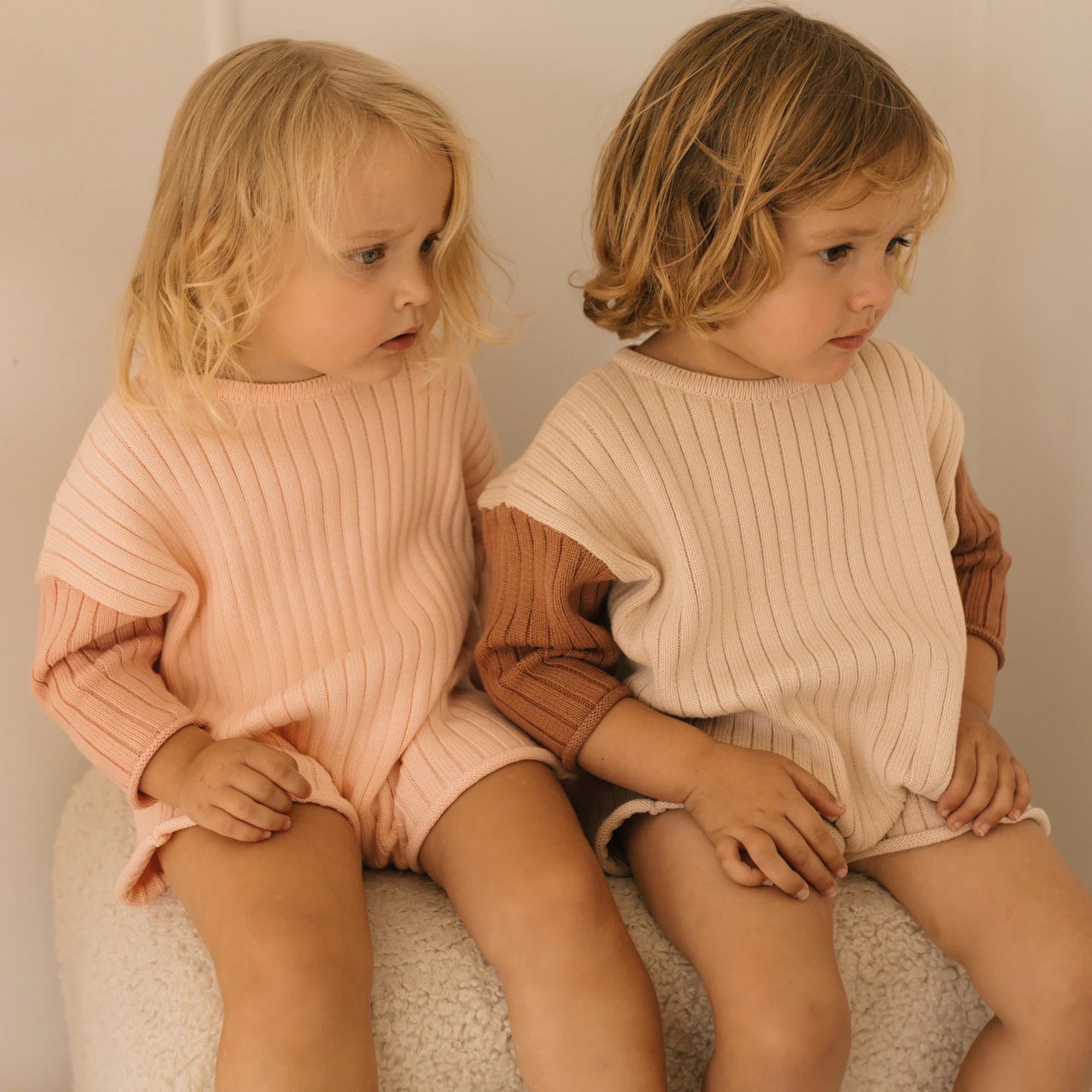 Two young children with blonde hair sit side by side on a fuzzy surface. They wear matching cotton knit rib outfits; the child on the left in a light pink romper and the child on the right in a stylish Golden Knit Romper Caramel Sundae from GOLDEN CHILDREN, crafted from neutral beige yarn with brown sleeves. Both look to their right.